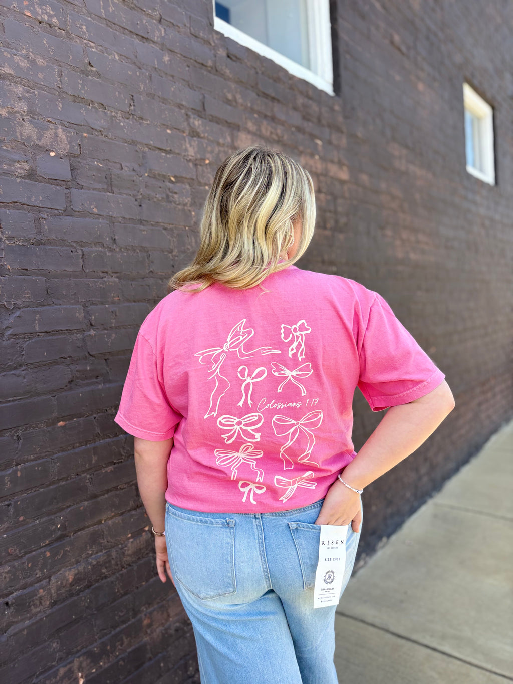 A girl with a pink bow shirt with denim jeans standing in front of a black brick wall