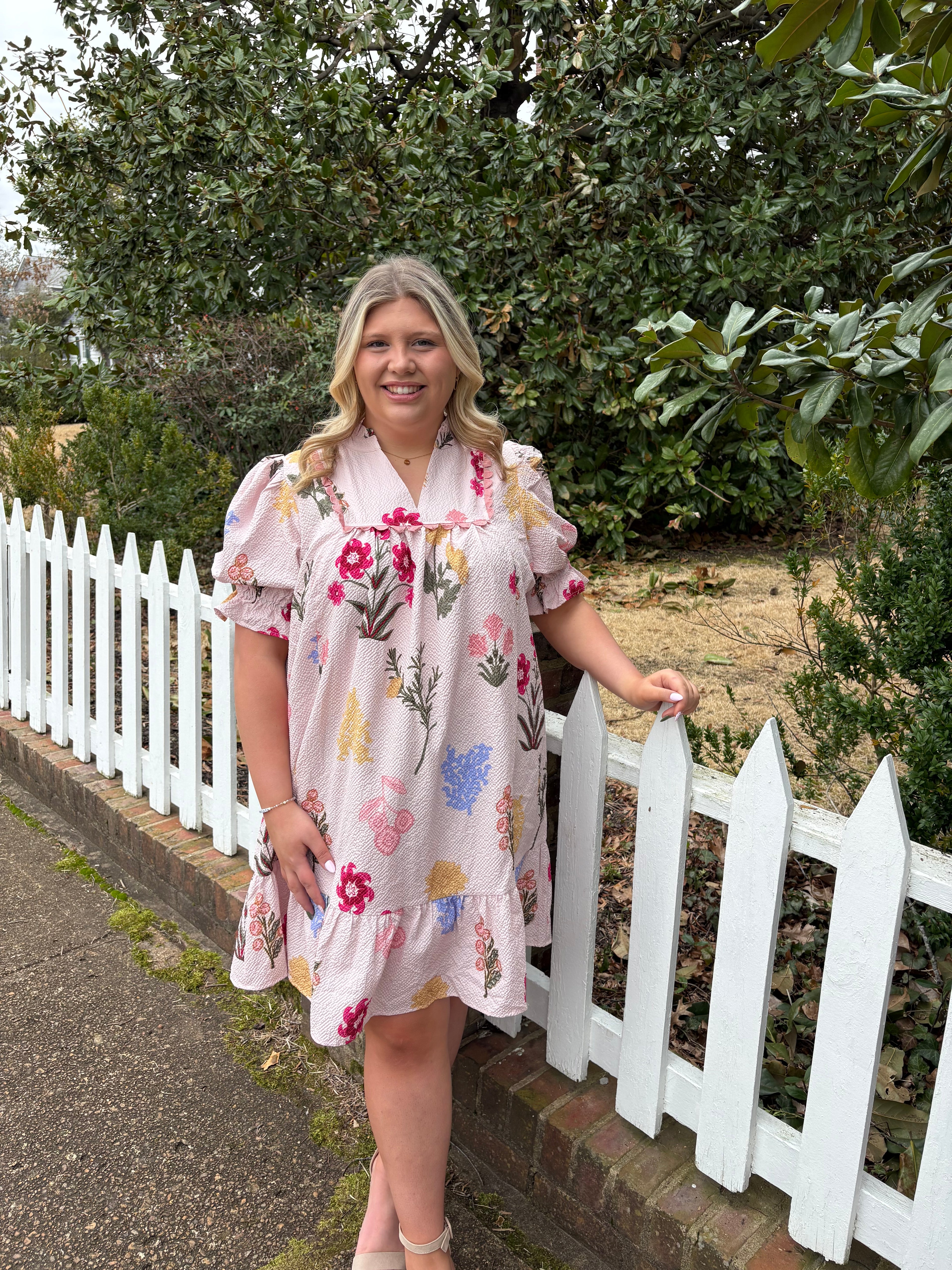 Woman in a floral dress standing next to a white picket fence with greenery in the background