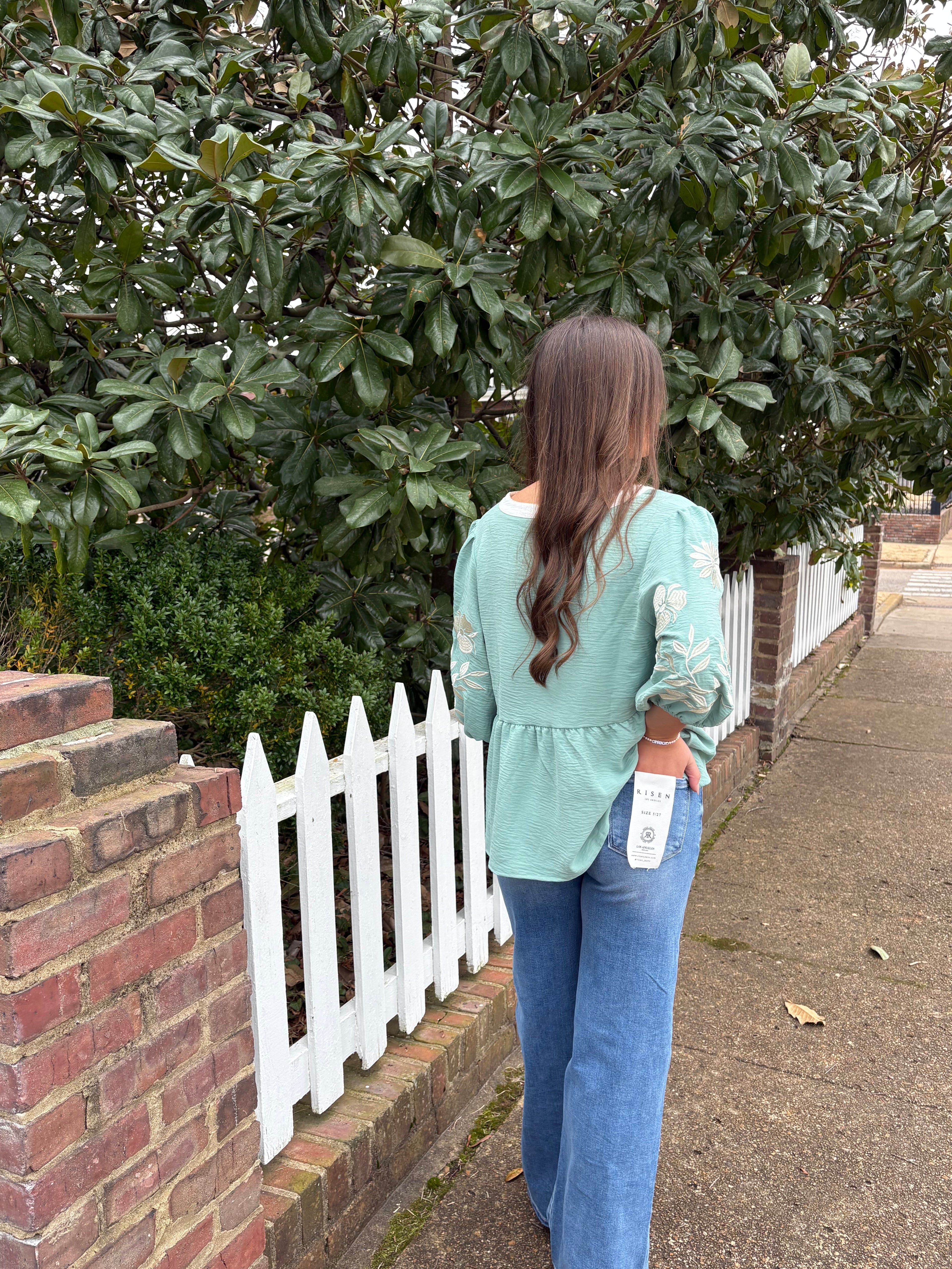 Person standing on a sidewalk next to a tree wearing a sage green blouse and denim blue jeans