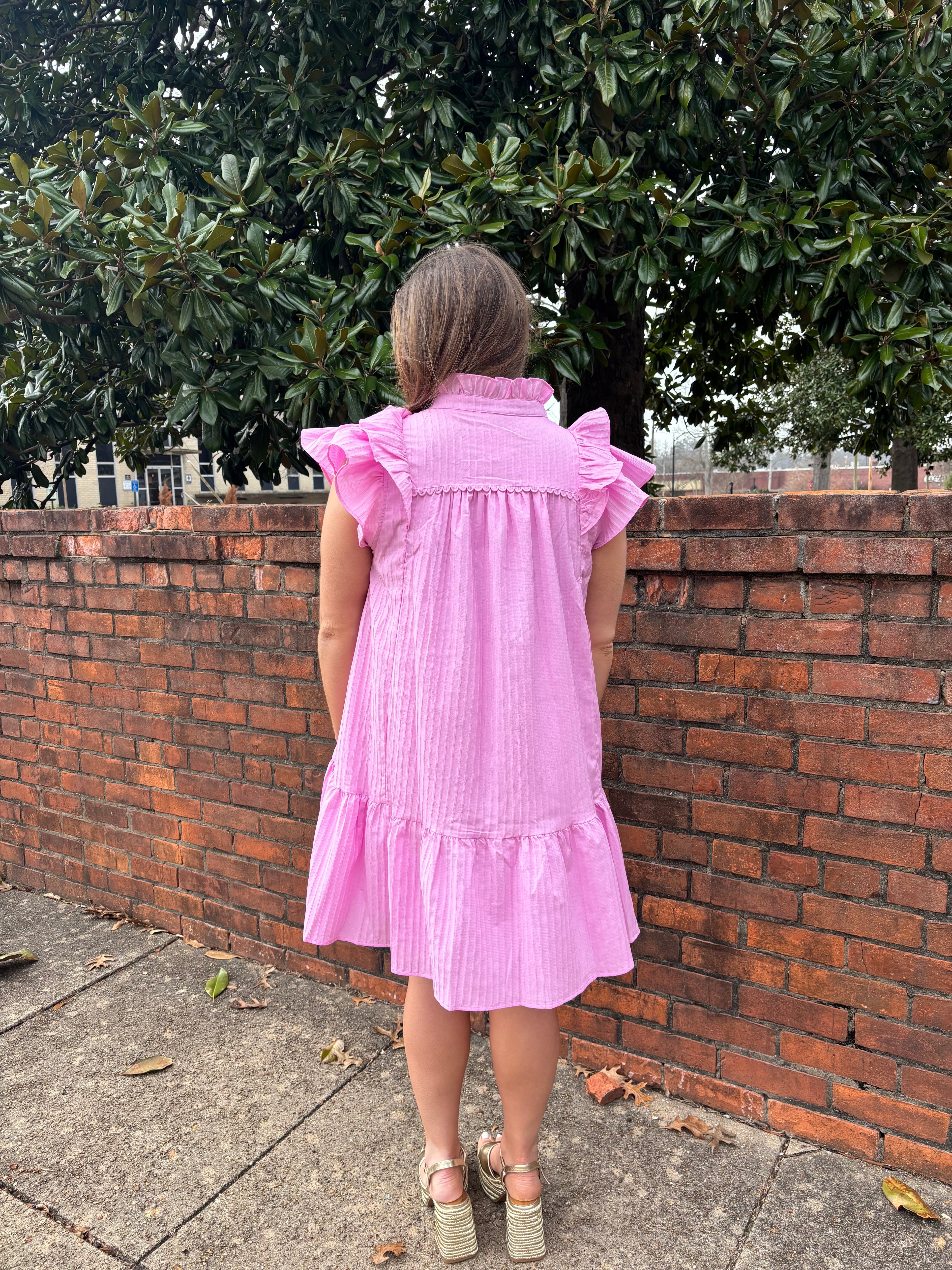 Person wearing a pink dress standing on a sidewalk with a brick wall and greenery in the background