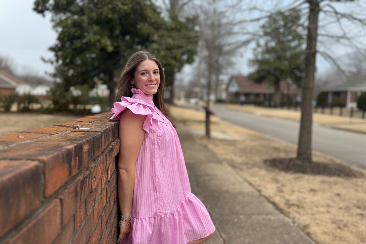 Person wearing a pink dress standing on a sidewalk with trees and buildings in the background