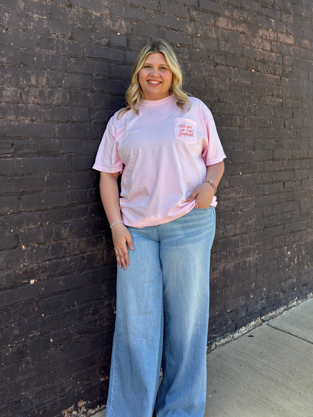 A girl in a light pink shirt wearing denim jeans standing in front of a black brick wall 