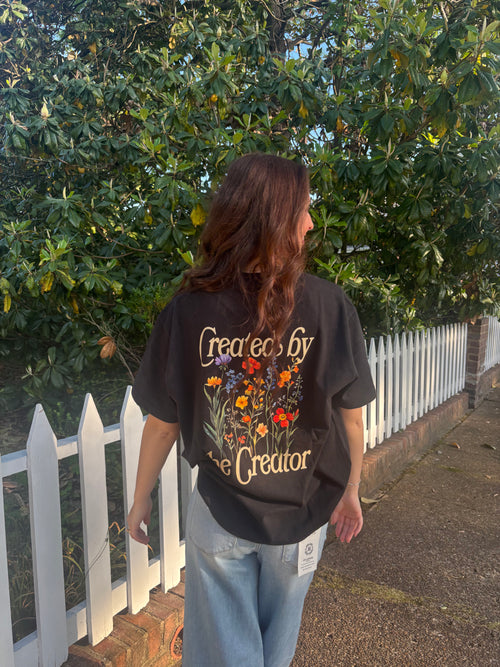 Person wearing a black t-shirt with floral designs and text, standing outdoors near a white picket fence.