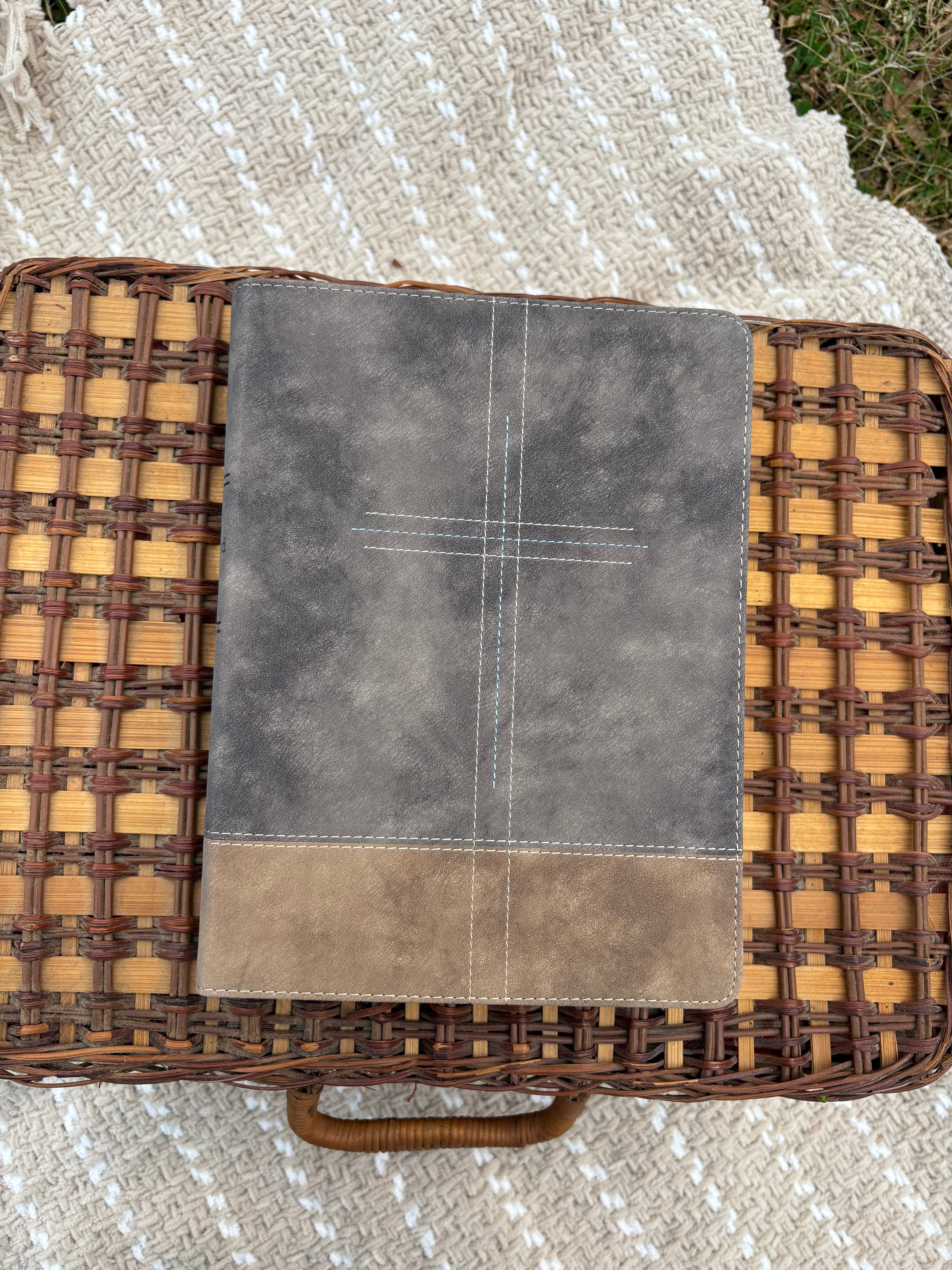Gray and brown bible on a woven basket laying on a textured beige blanket outdoors