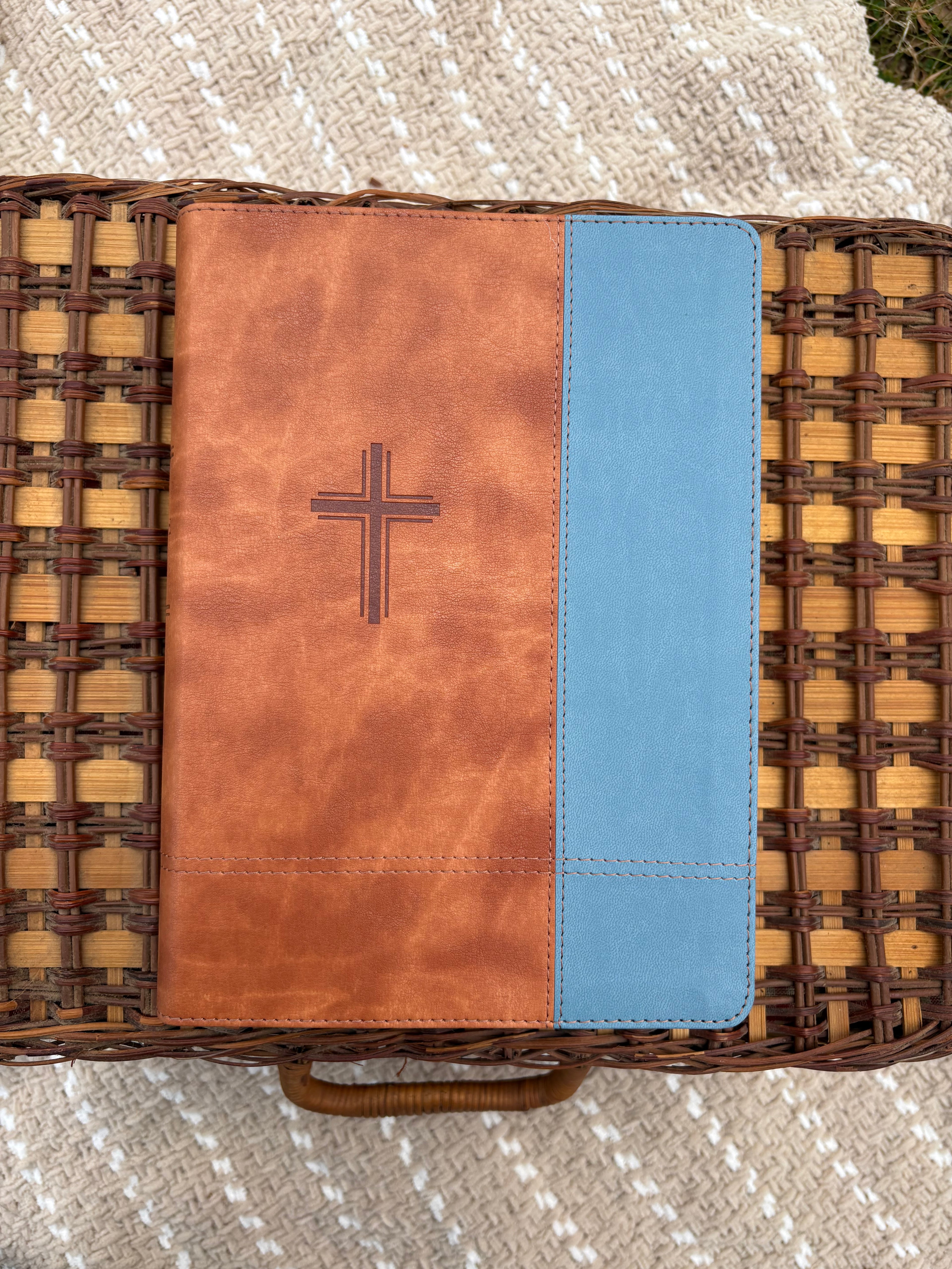 Brown leather bible with blue top section on a woven basket on beige blanket outdoors