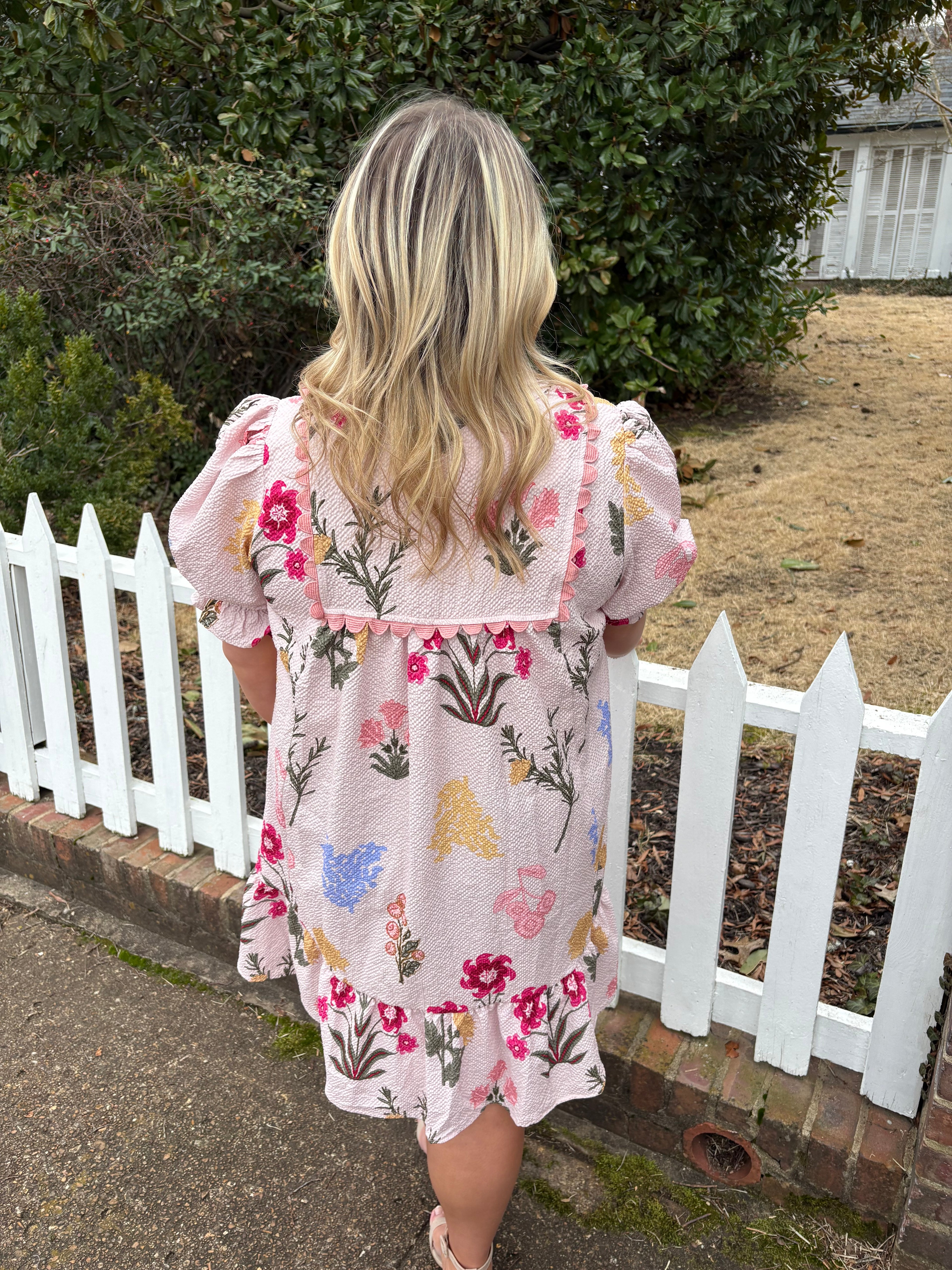 Person wearing a floral dress standing next tia white picket fence outdoors.