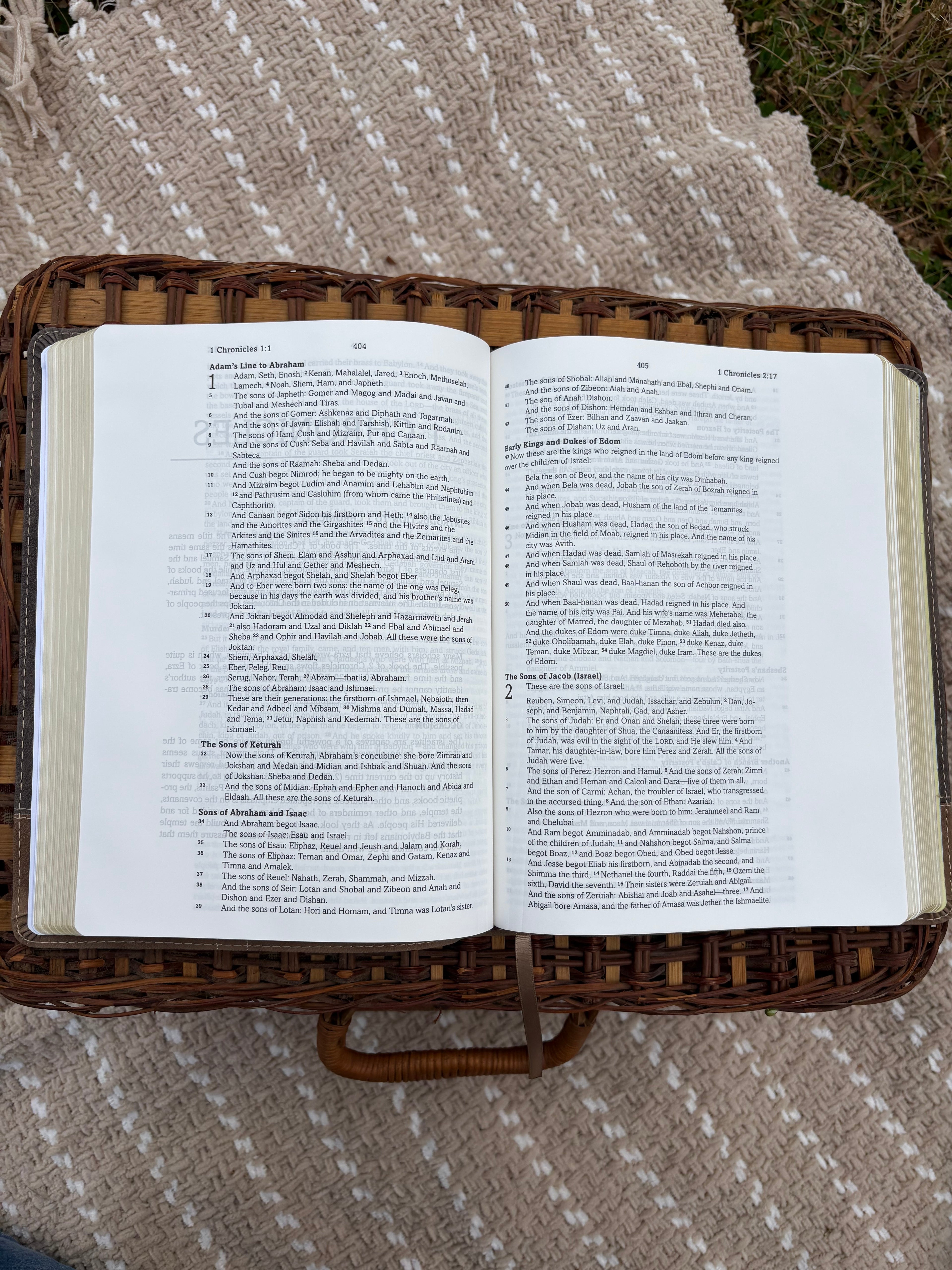 Open bible on a wicker basket with text pages, laying on a beige blanket outdoors.