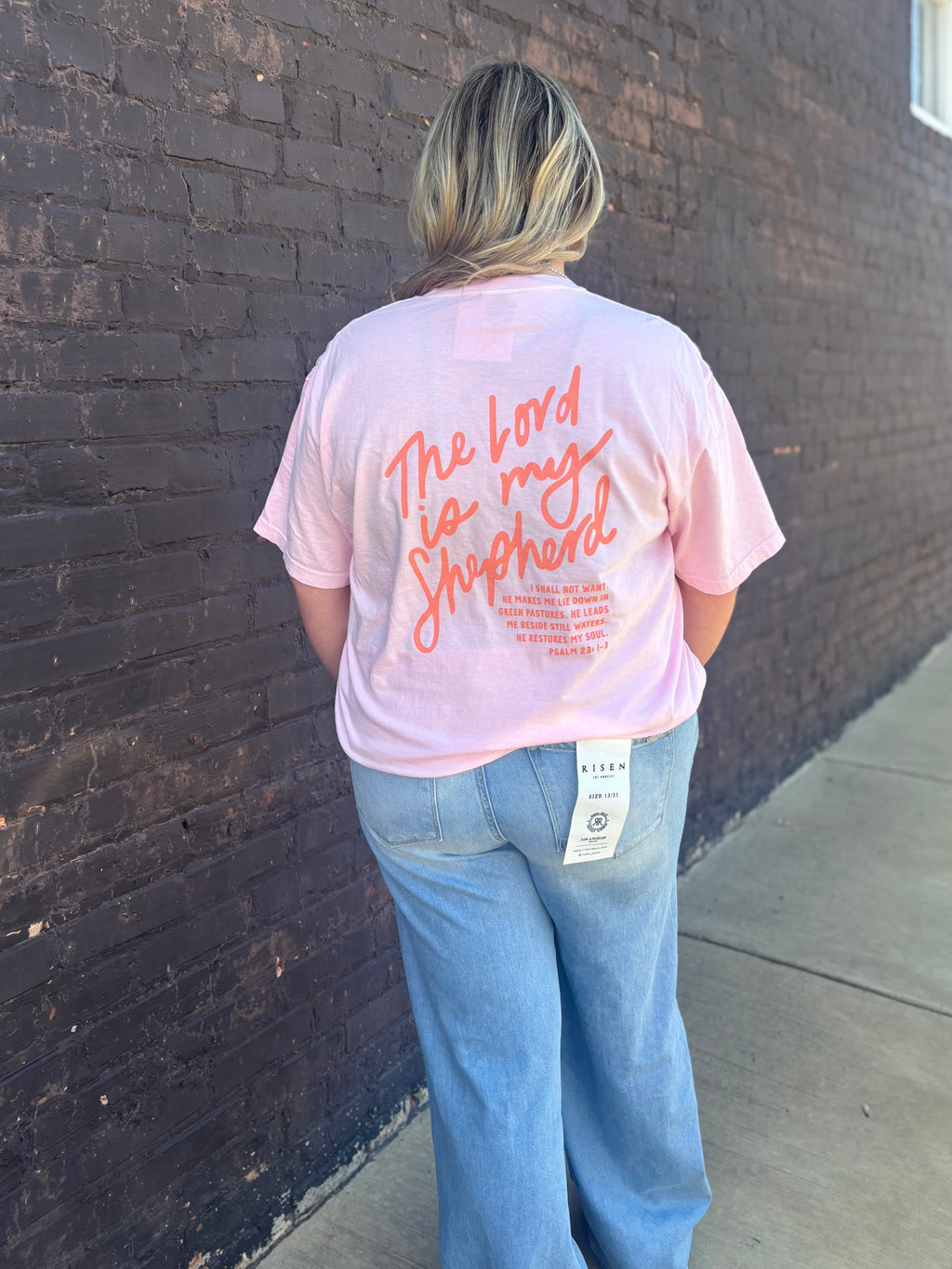 A girl in a light pink shirt wearing denim jeans standing in front of a black brick wall 