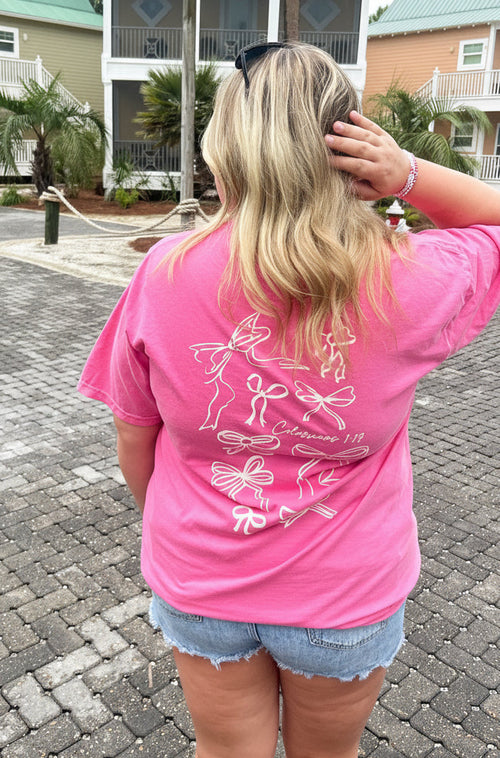 Person wearing a pink t-shirt with a design on the back, standing on a brick driveway with houses in the background.