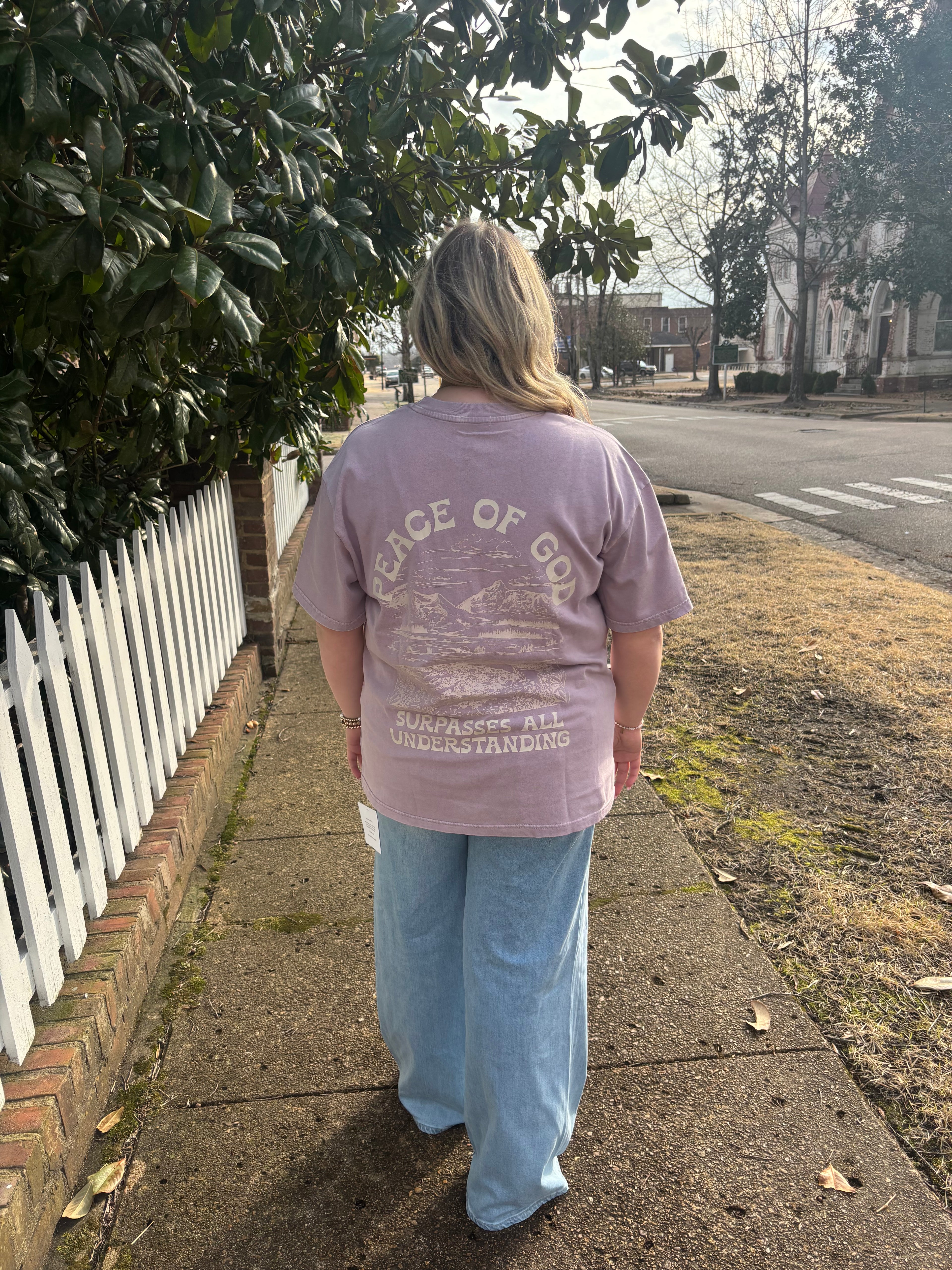 Person wearing a light purple t-shirt with text, standing on a sidewalk next to a bench.