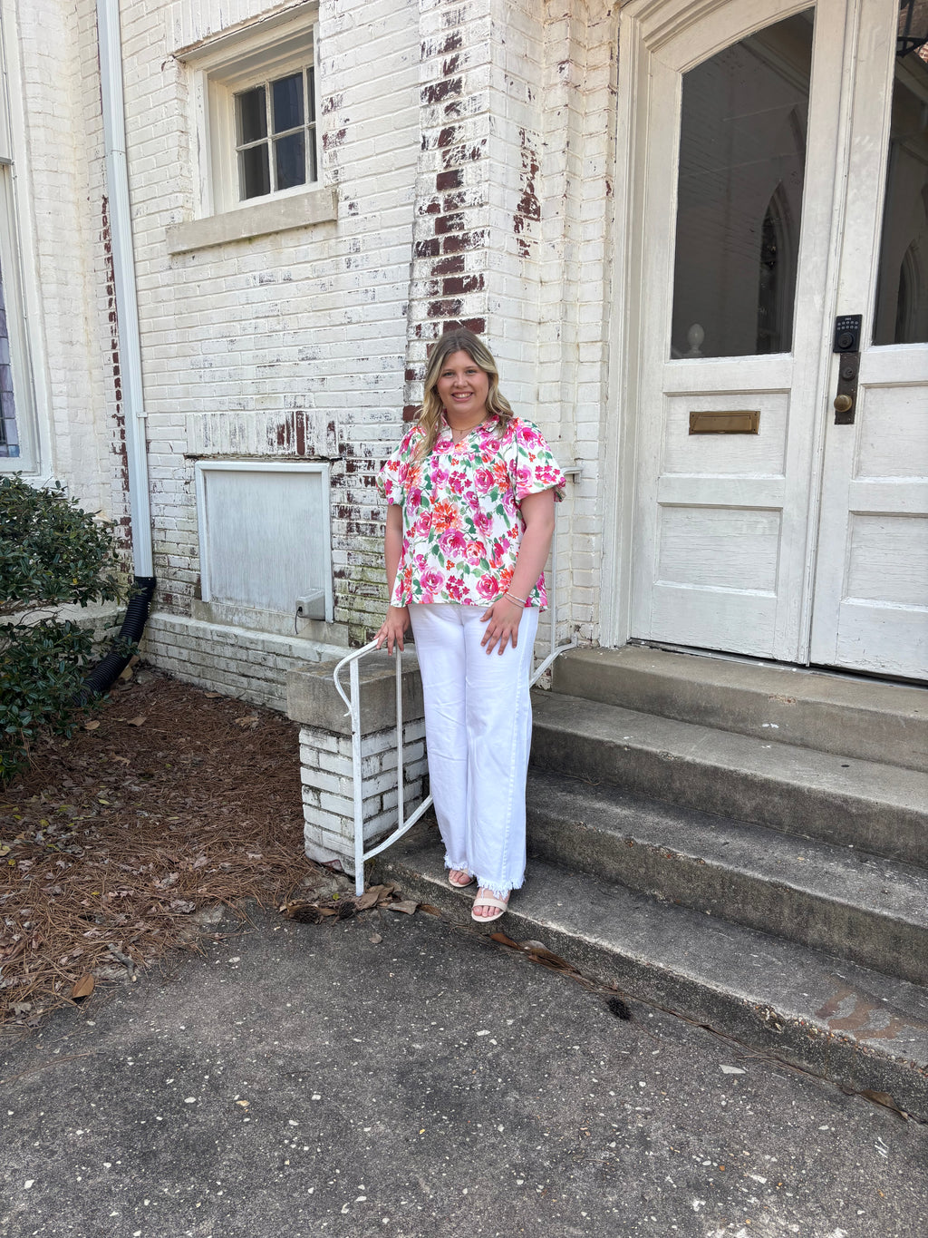 Person standing on a porch with a floral shirt and white pants, leaning against a railing.