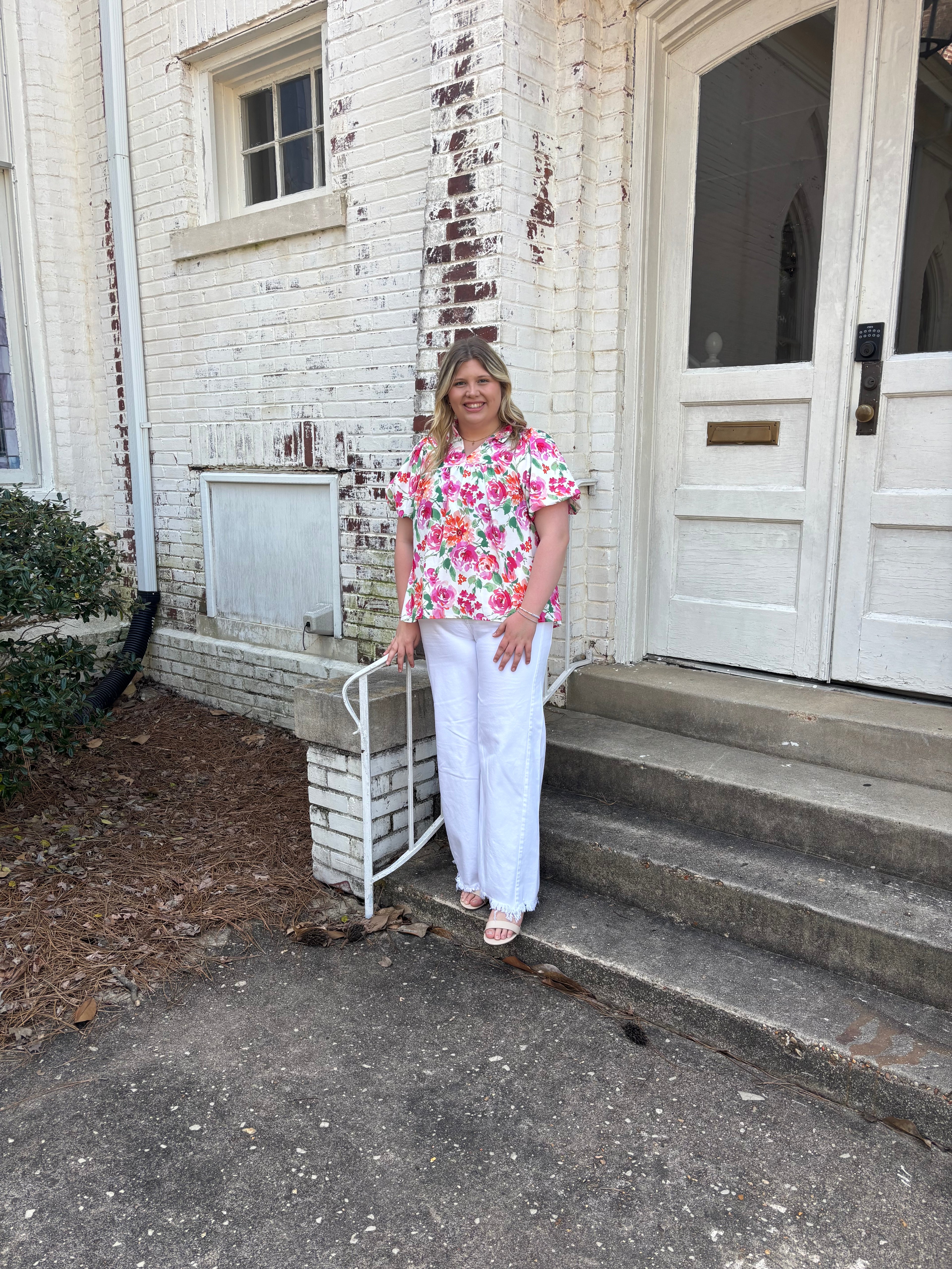 Person standing on a porch with a floral shirt and white pants, leaning against a railing.