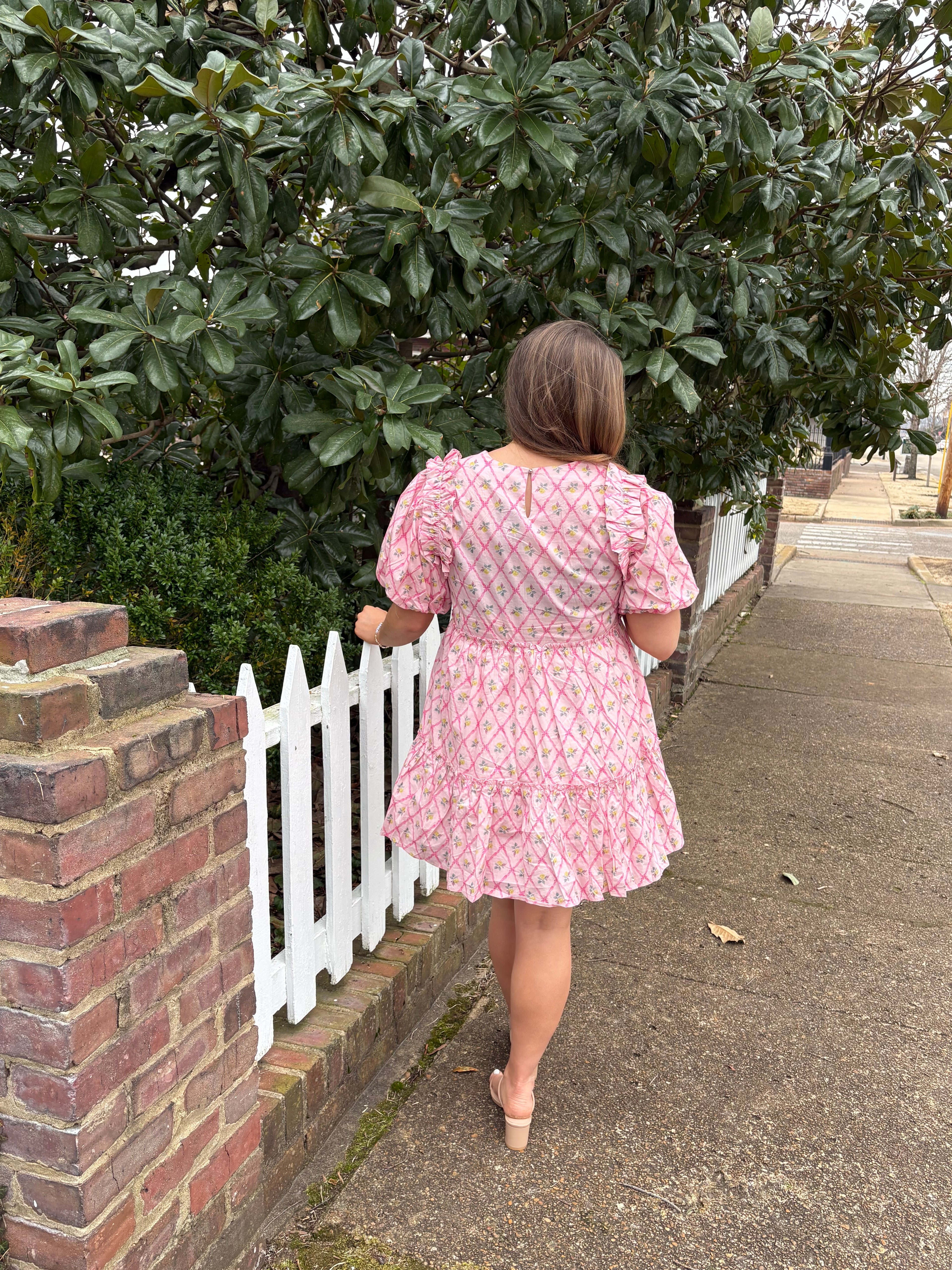 Person in a pink dress standing beside a white picket fence with greenery and a sidewalk in the background