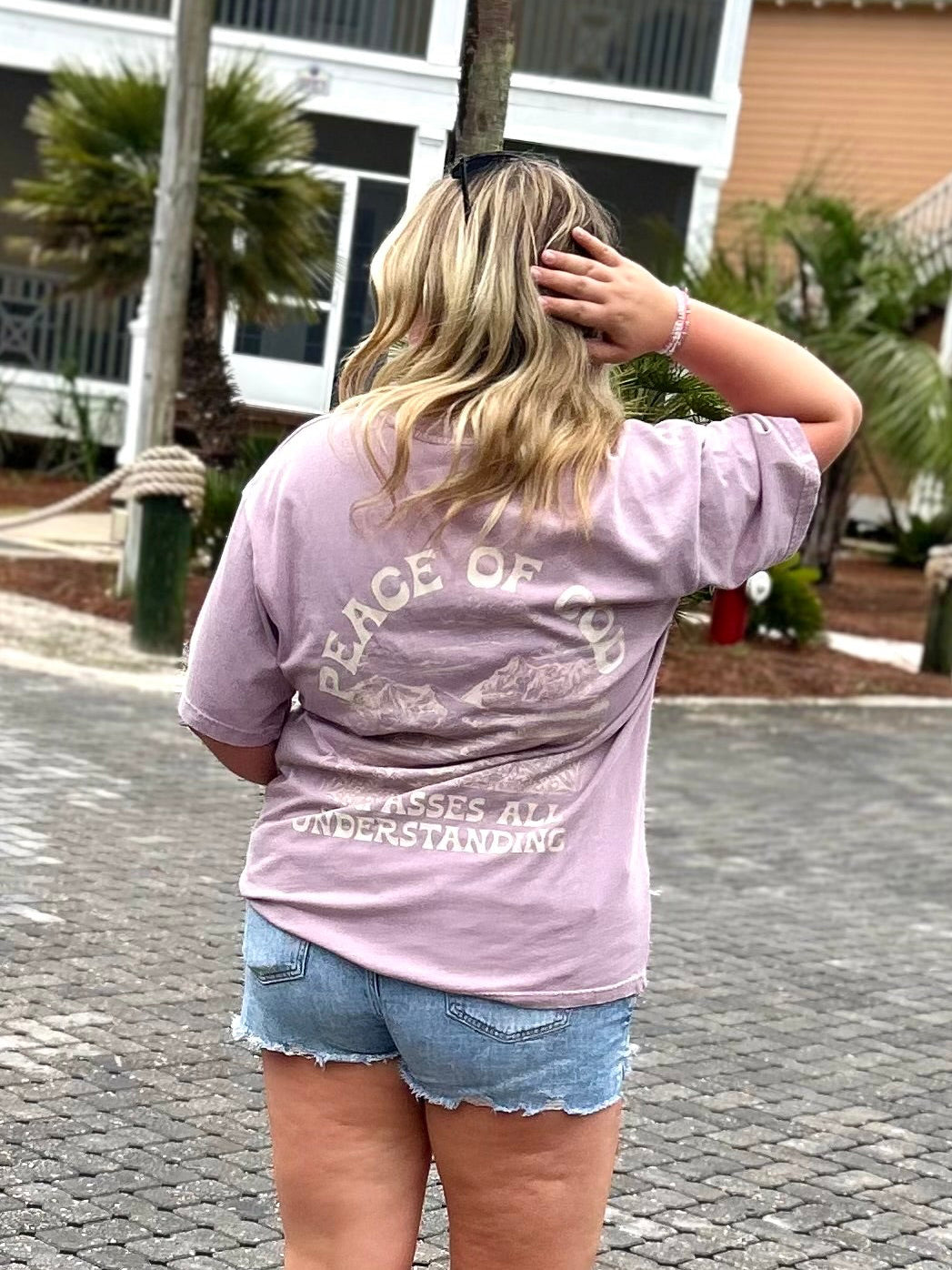 Person wearing a light purple t-shirt with text and graphics, standing on a paved area with palm trees and buildings in the background.