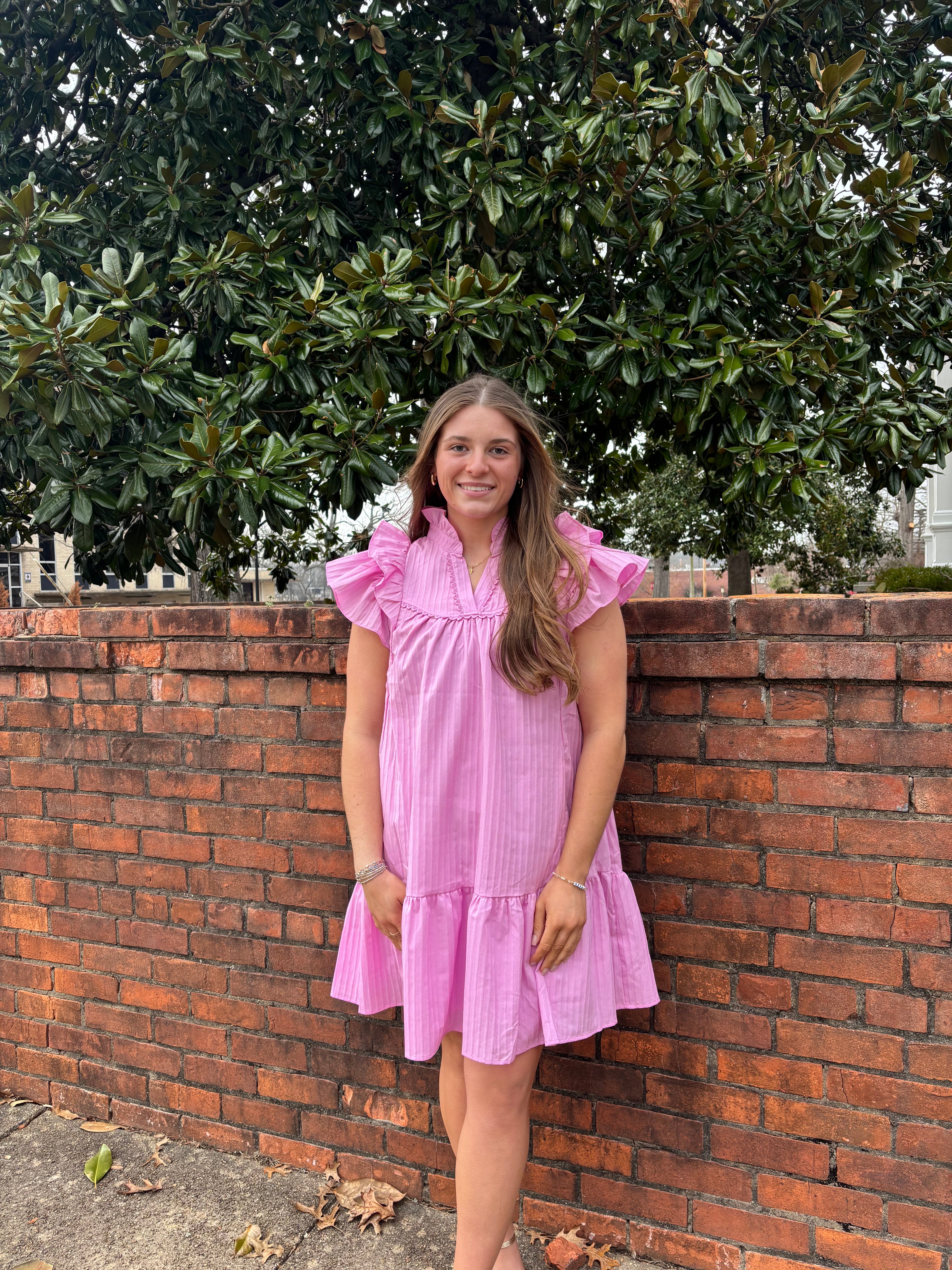 Woman in a pink dress standing outdoors next to a brick wall and tree