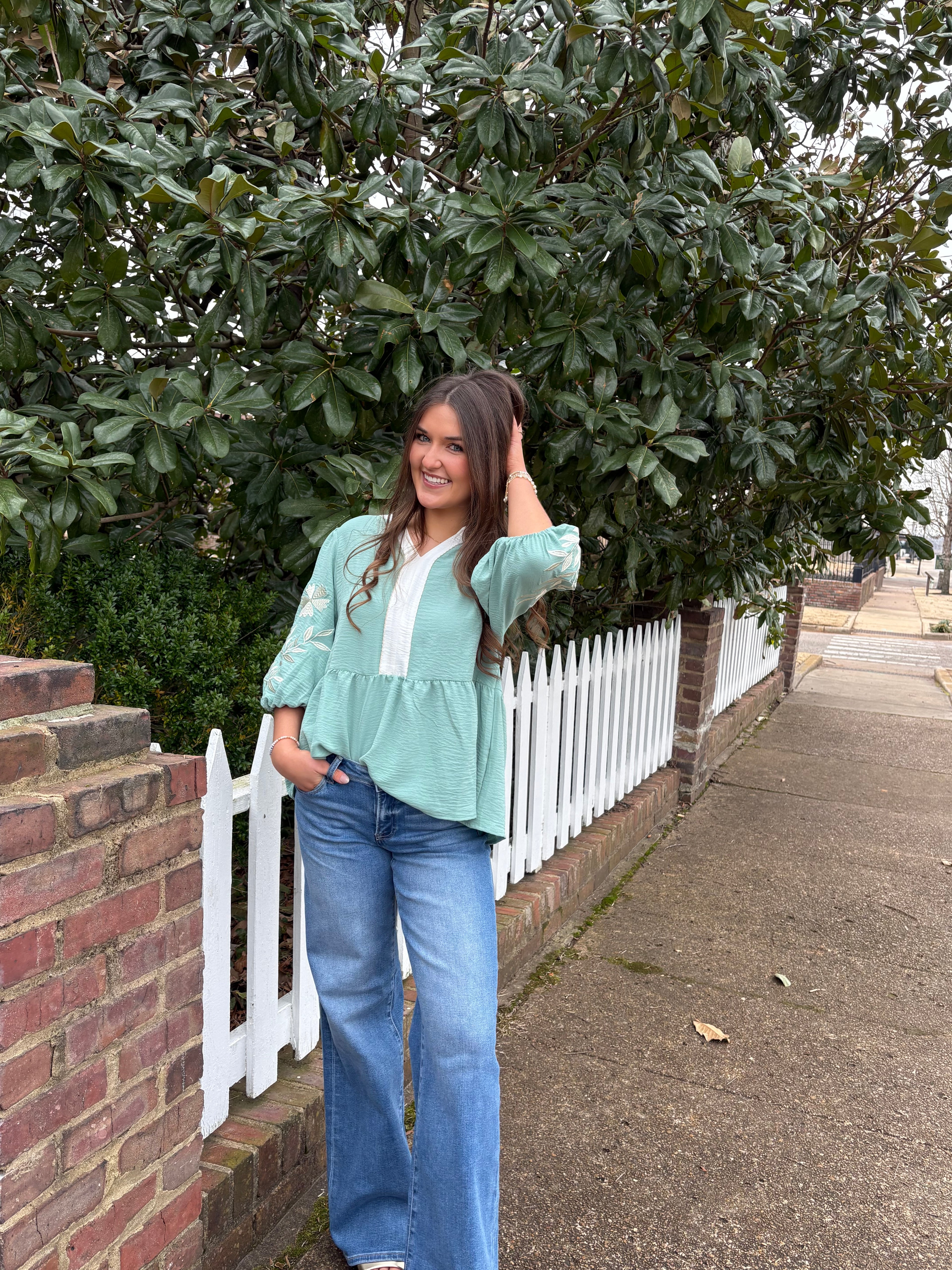 Person standing on a sidewalk next to a tree wearing a sage green blouse and denim blue jeans