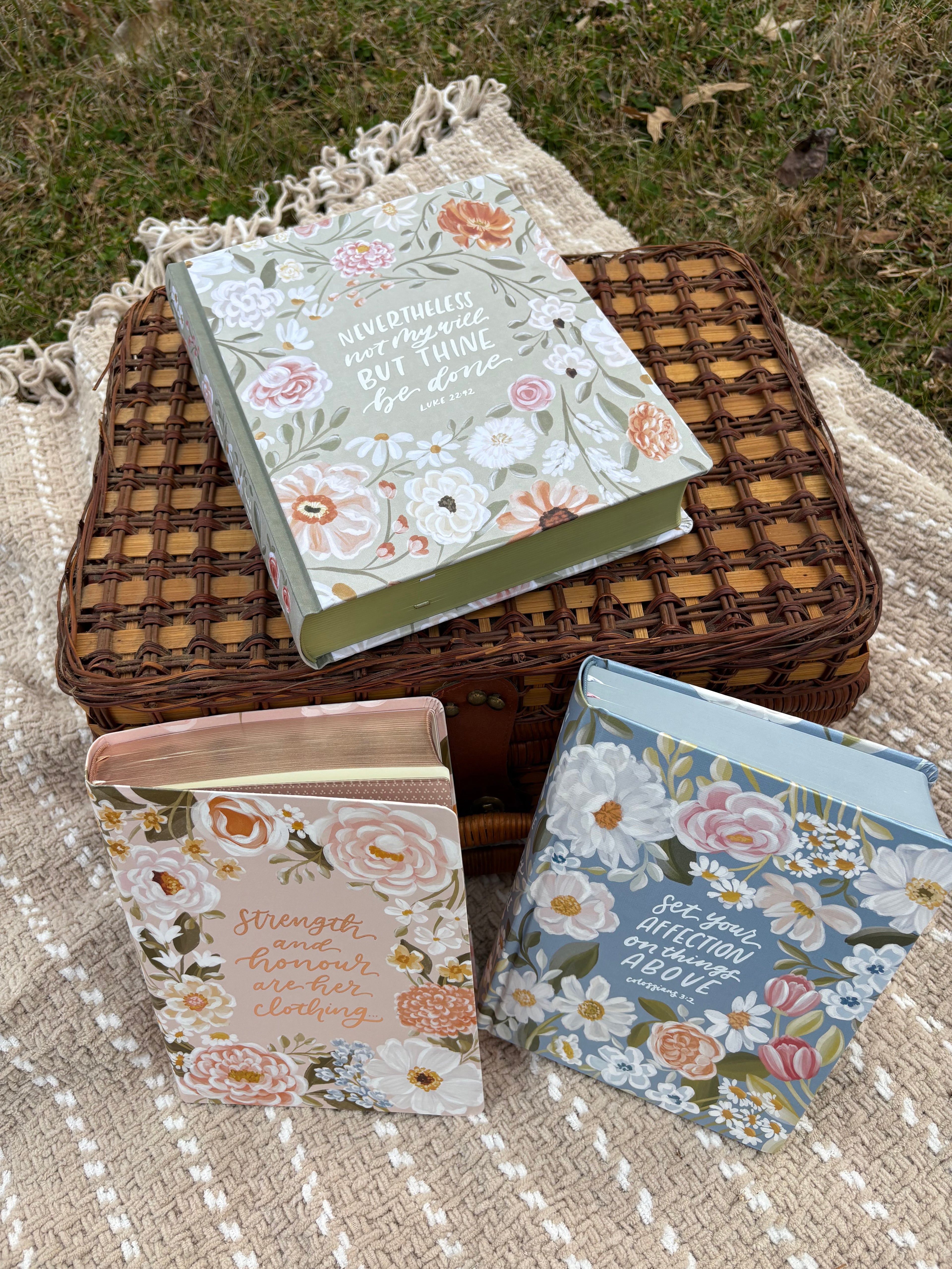 Three floral-patterned bibles, a textured beige blanket with a woven basket. on the ground