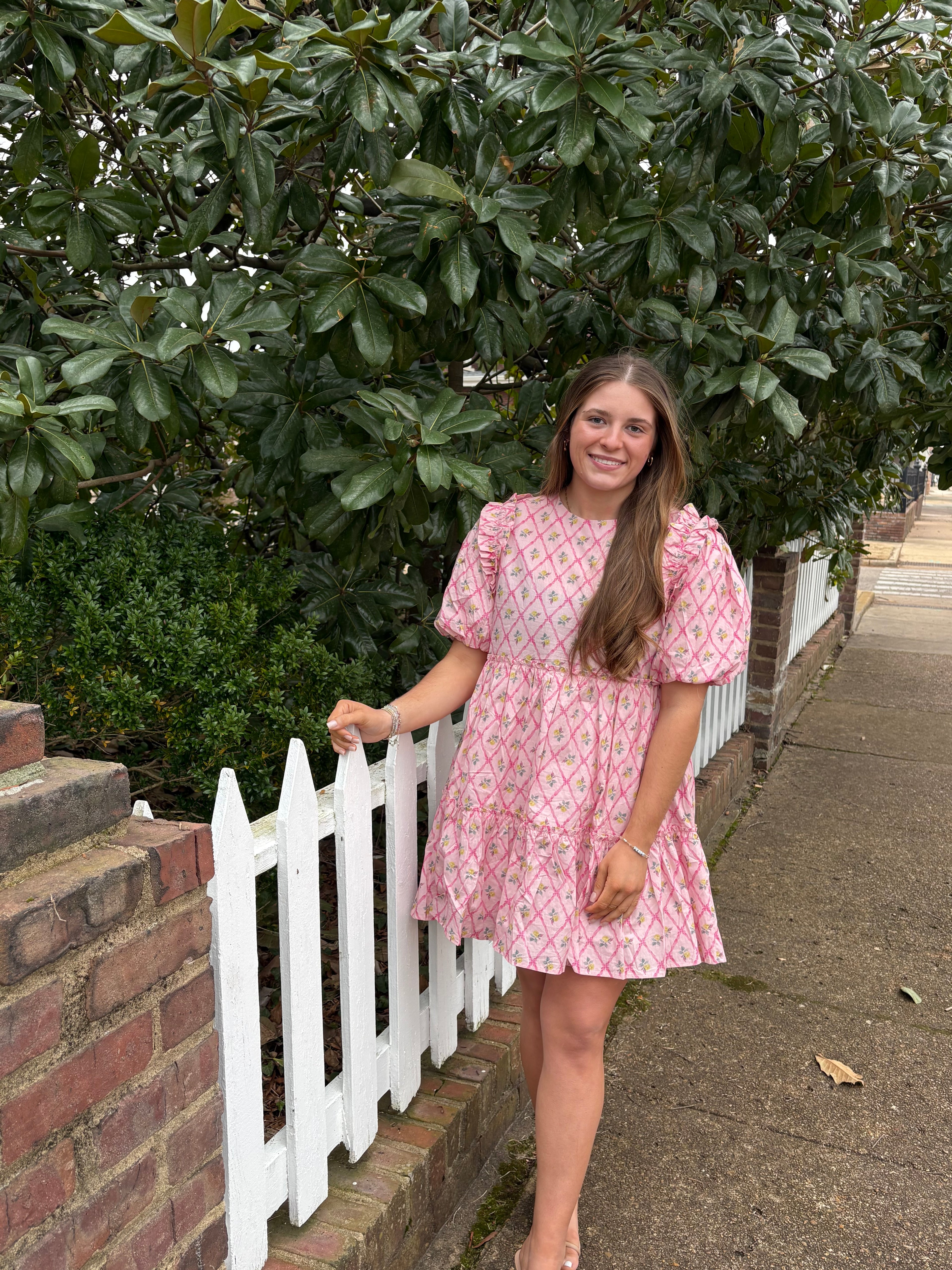 Person in a pink dress standing beside a white picket fence with greenery and a sidewalk in the background