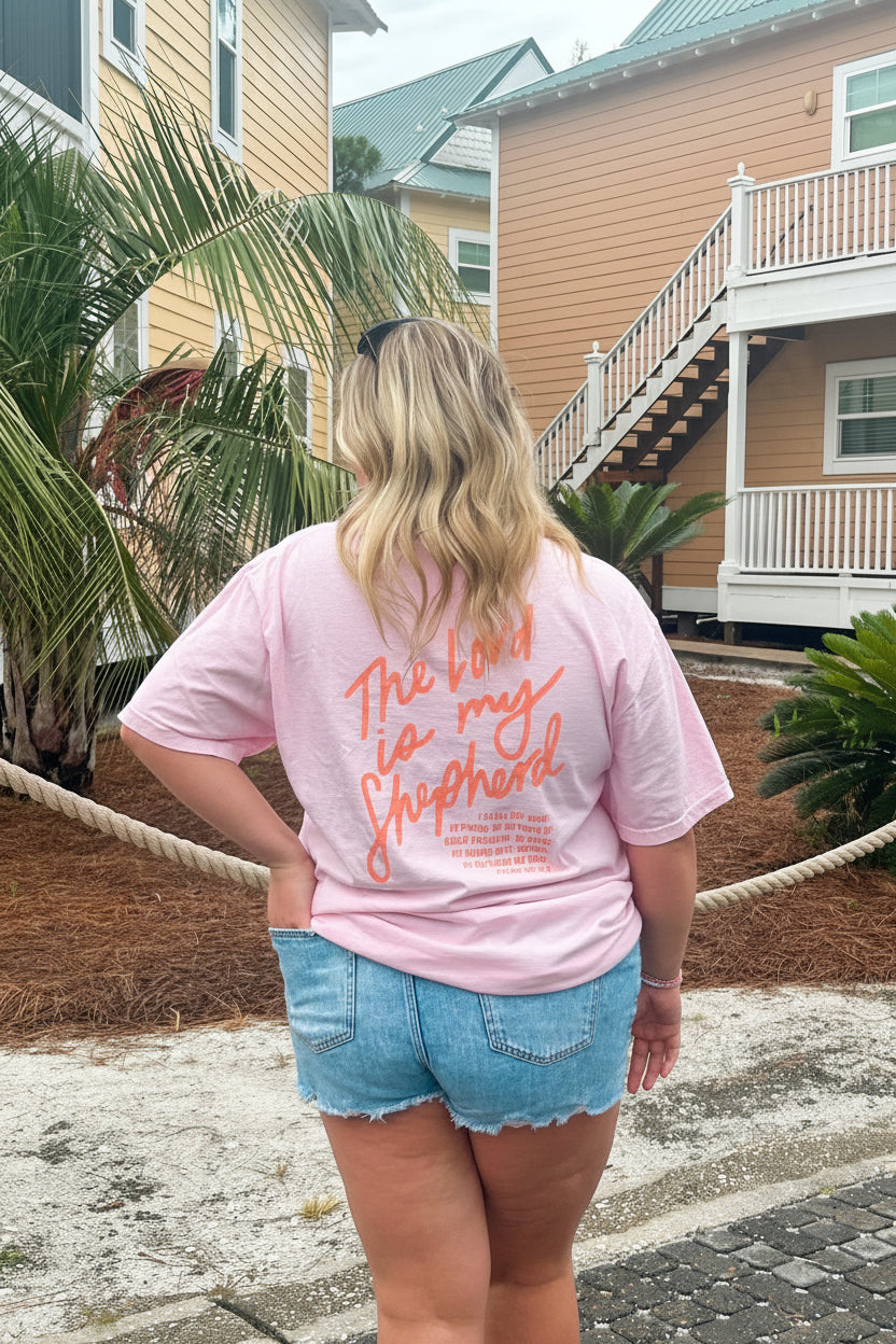 Person wearing a pink t-shirt with text and denim shorts standing outdoors.