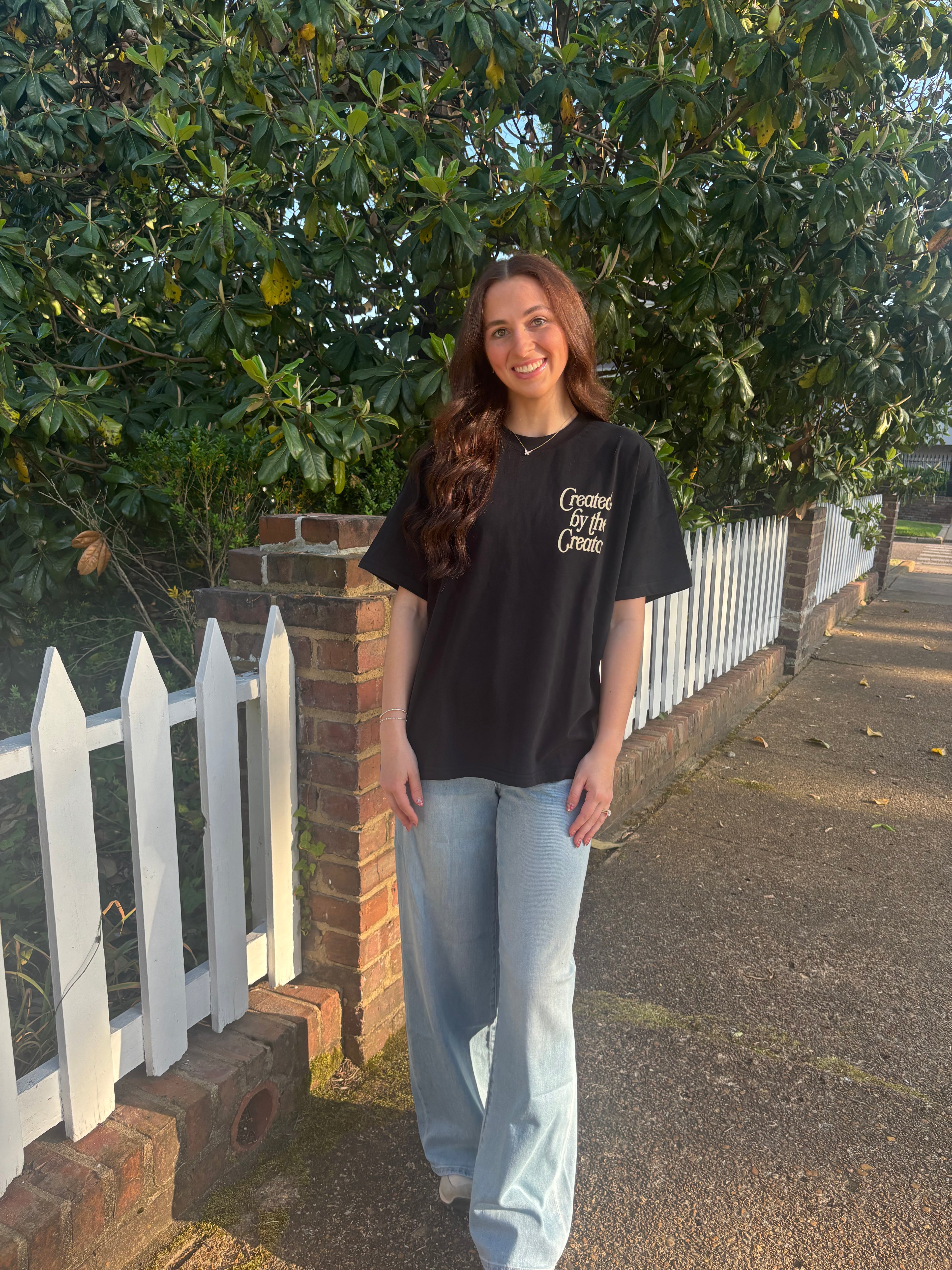 Person wearing a black t-shirt with text, standing outdoors near a white picket fence and greenery.