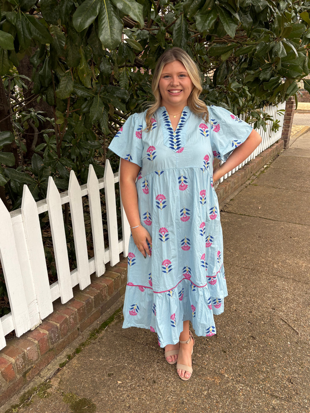 Woman in a light blue dress with floral patterns standing on a sidewalk next to a white picket fence.