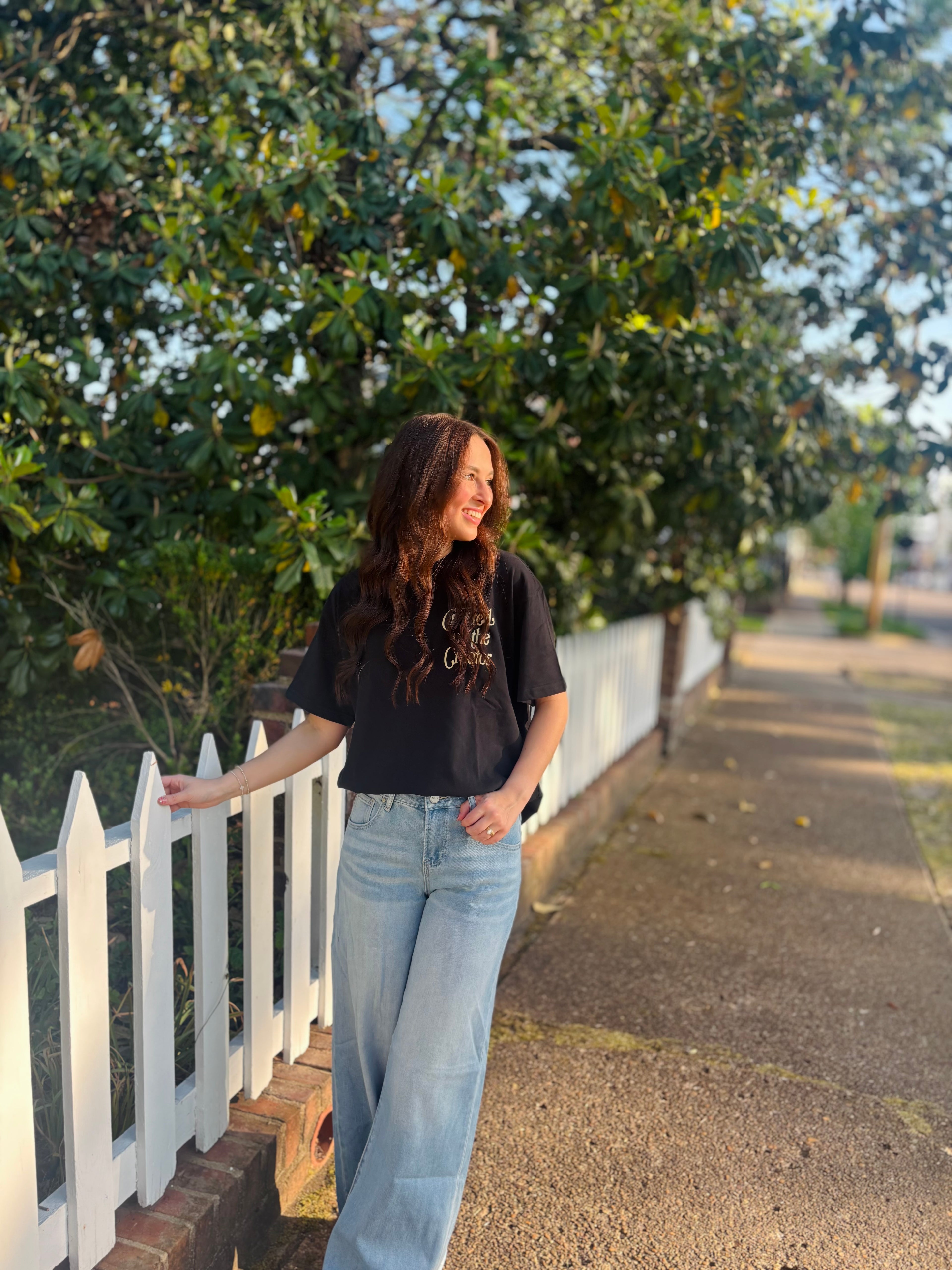 Woman standing on a sidewalk next to a white picket fence with trees in the background
