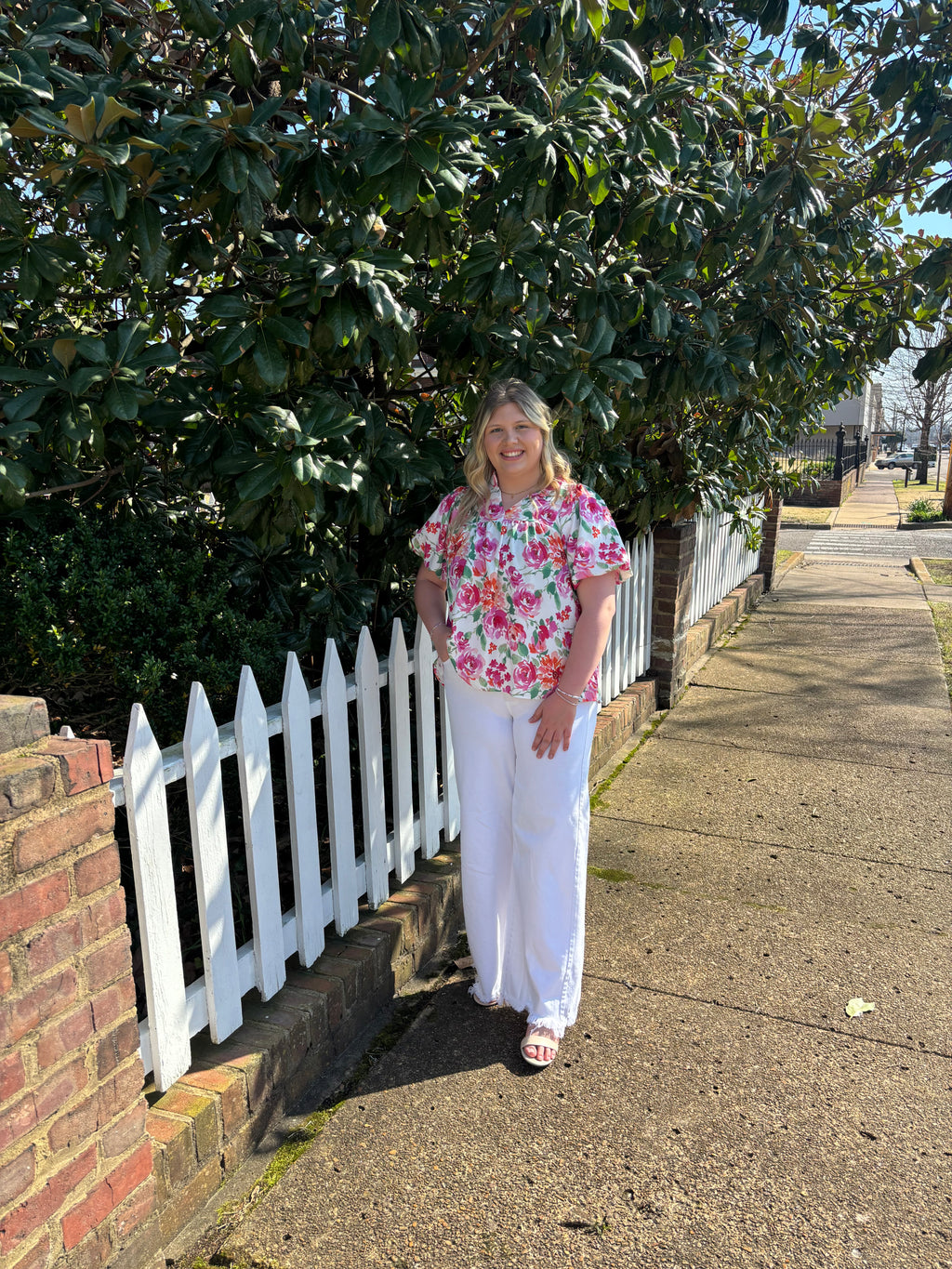Woman standing on a sidewalk next to a white picket fence with greenery in the background