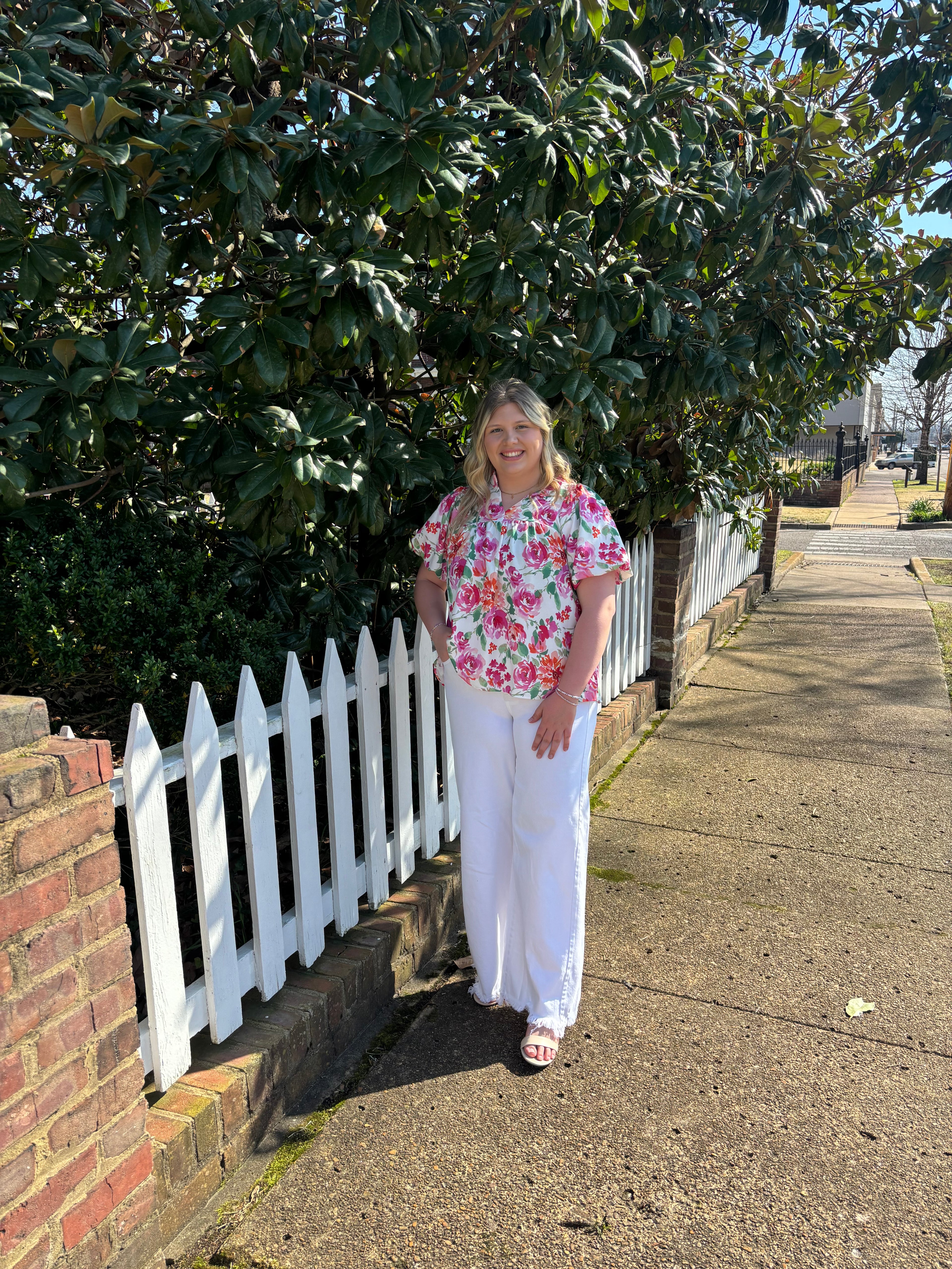 Woman standing on a sidewalk next to a white picket fence with greenery in the background