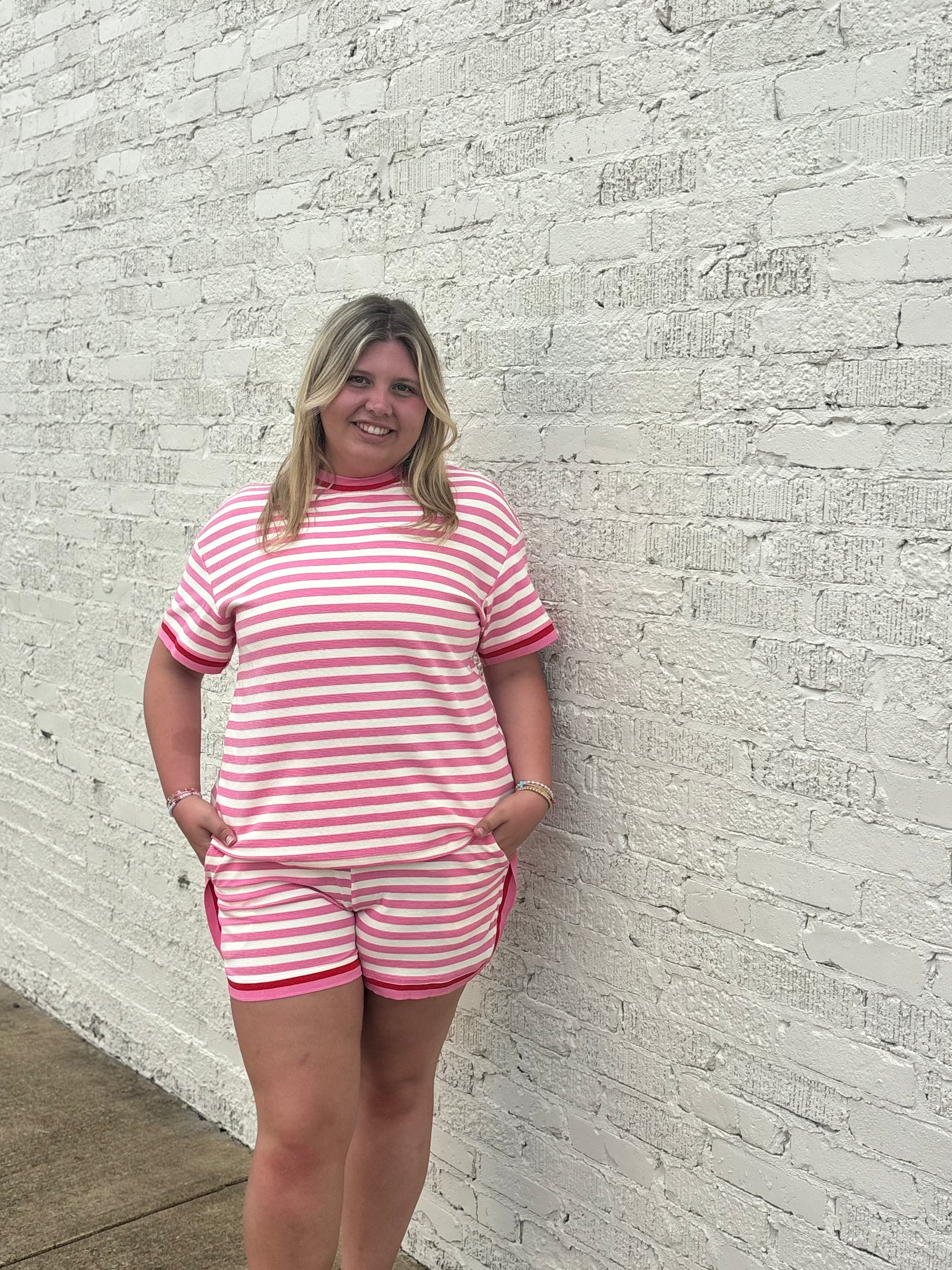Woman wearing a pink and white striped outfit standing against a light gray brick wall.