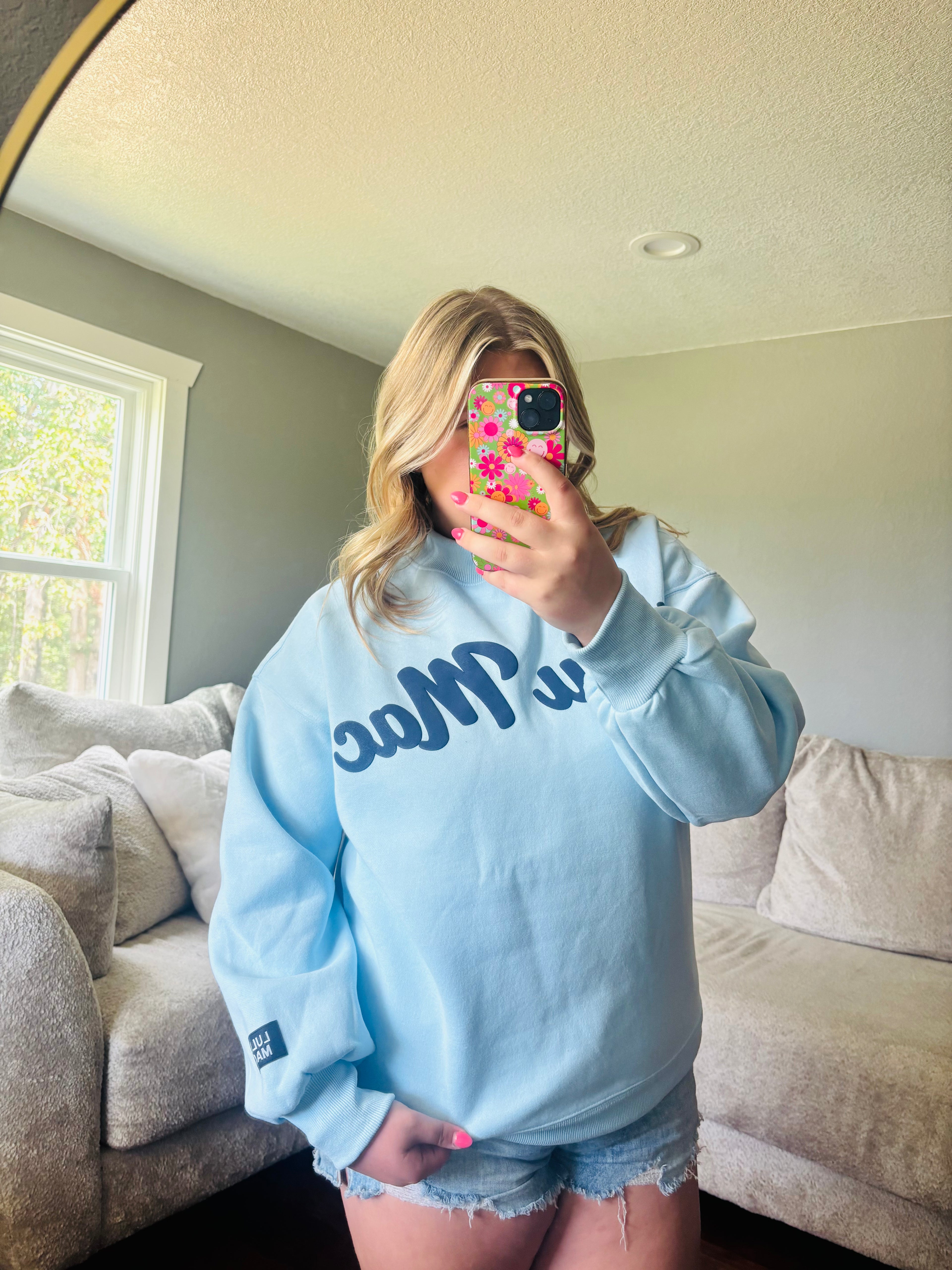 A woman takes a mirror selfie in a living room, wearing an oversized light blue sweatshirt with dark blue script lettering across the chest and denim shorts.