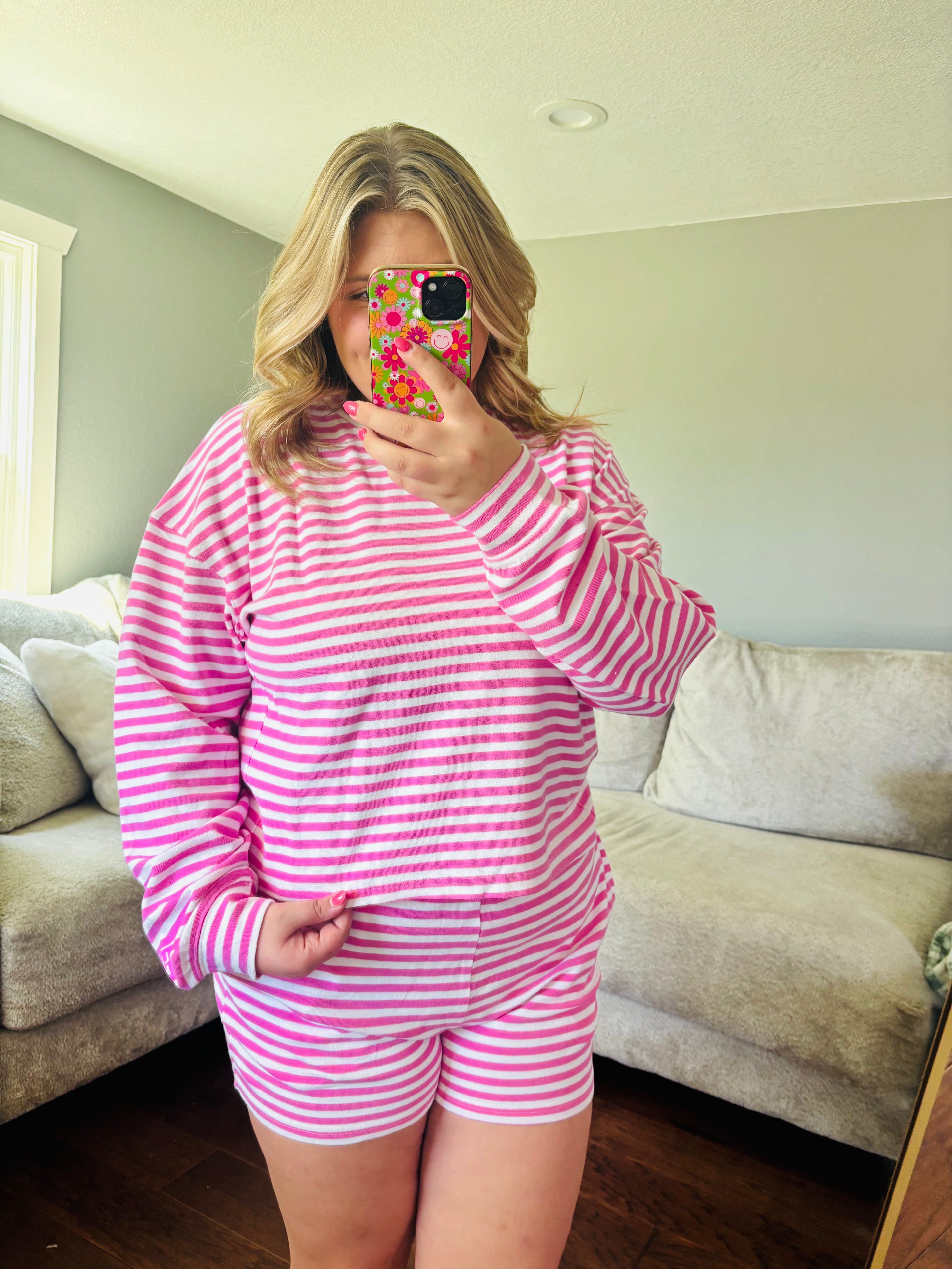 A woman takes a mirror selfie wearing a pink and white striped long-sleeve top and matching shorts in a living room setting.
