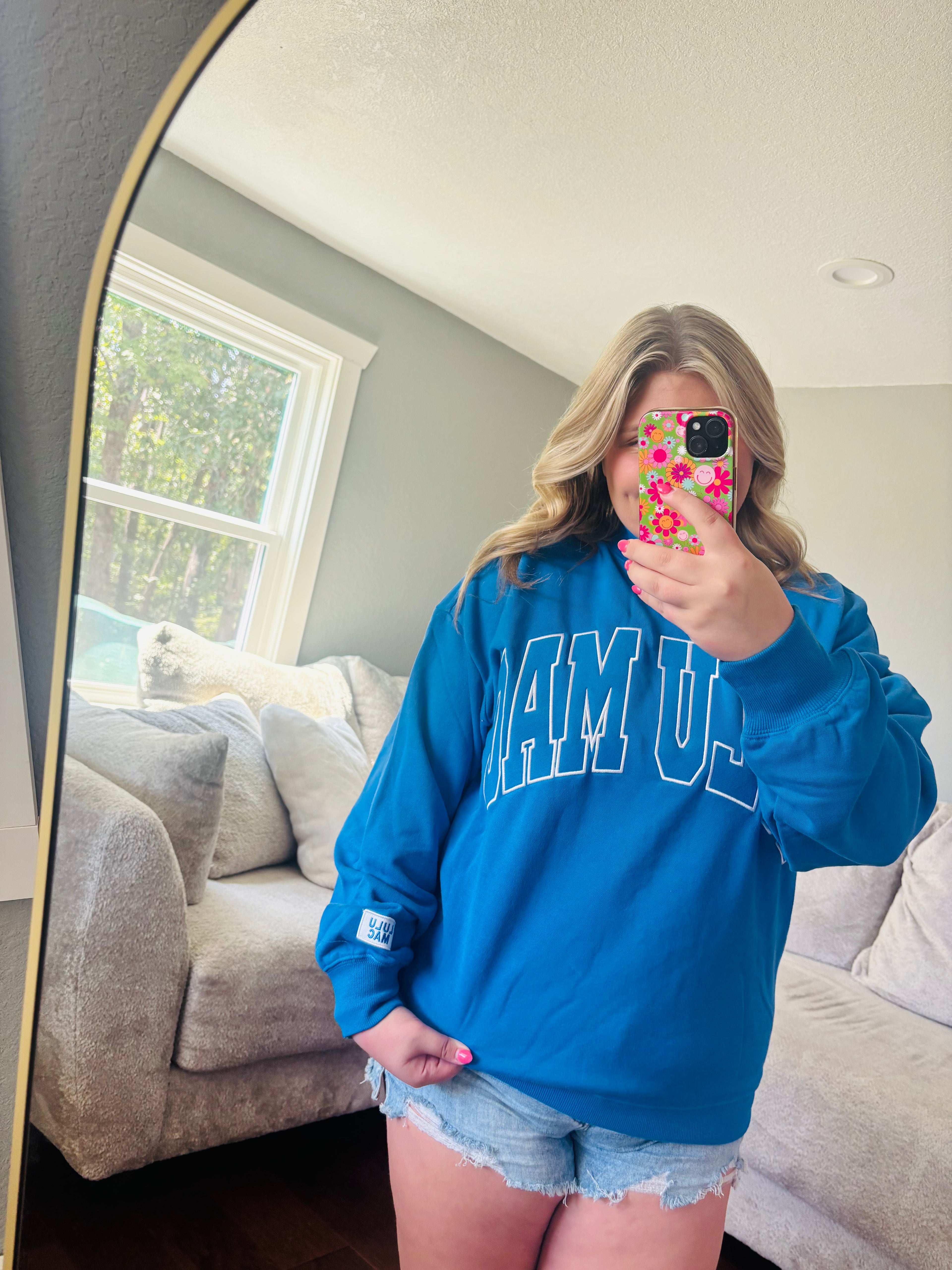 A woman takes a mirror selfie in a living room, wearing a bright blue sweatshirt with 'LULU MAC' printed in white arched letters across the chest, paired with denim shorts.