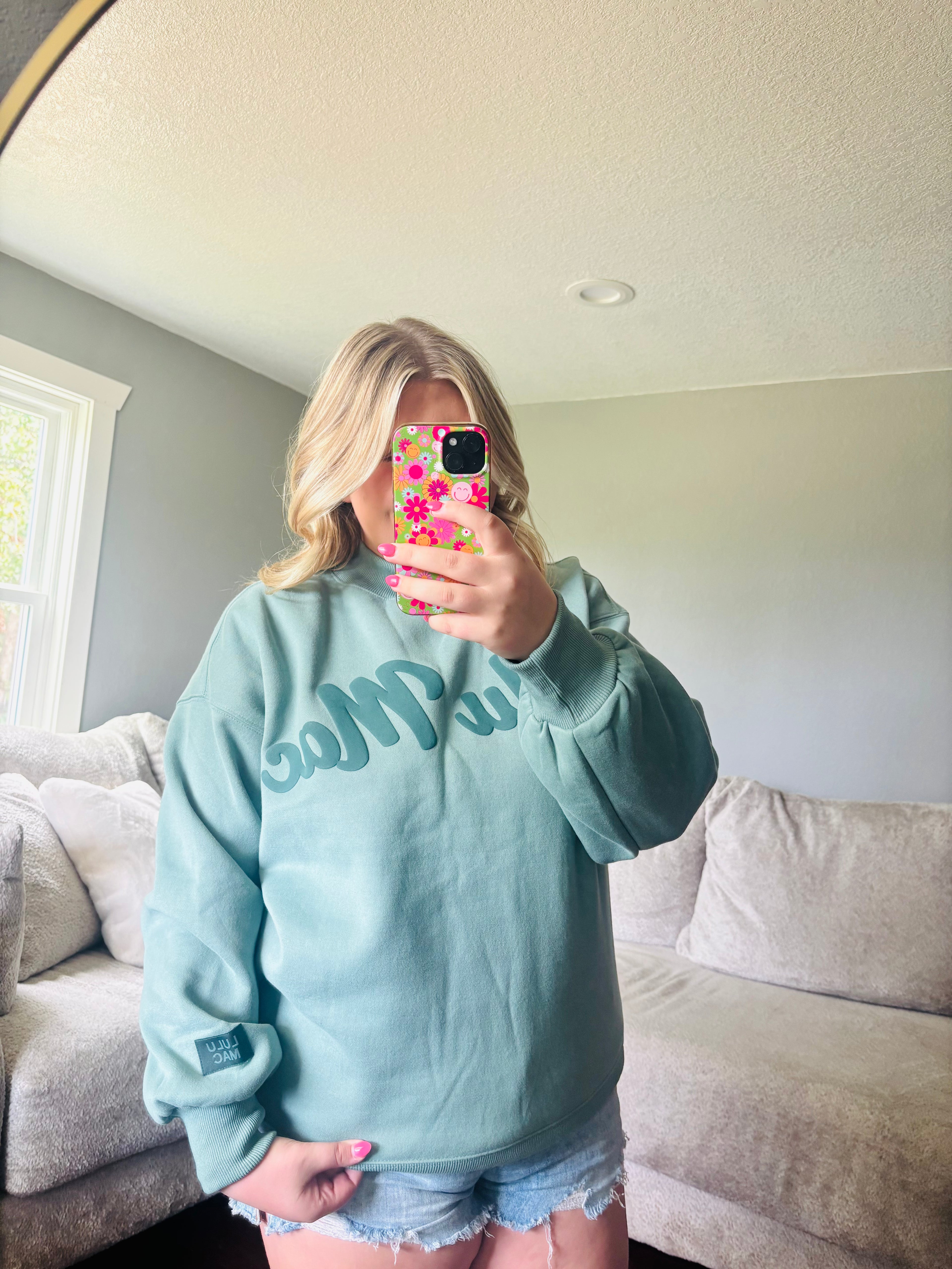 A woman takes a mirror selfie in a living room, wearing a light blue sweatshirt with 'LULU MAC' written across the chest in a darker blue script, paired with denim shorts.