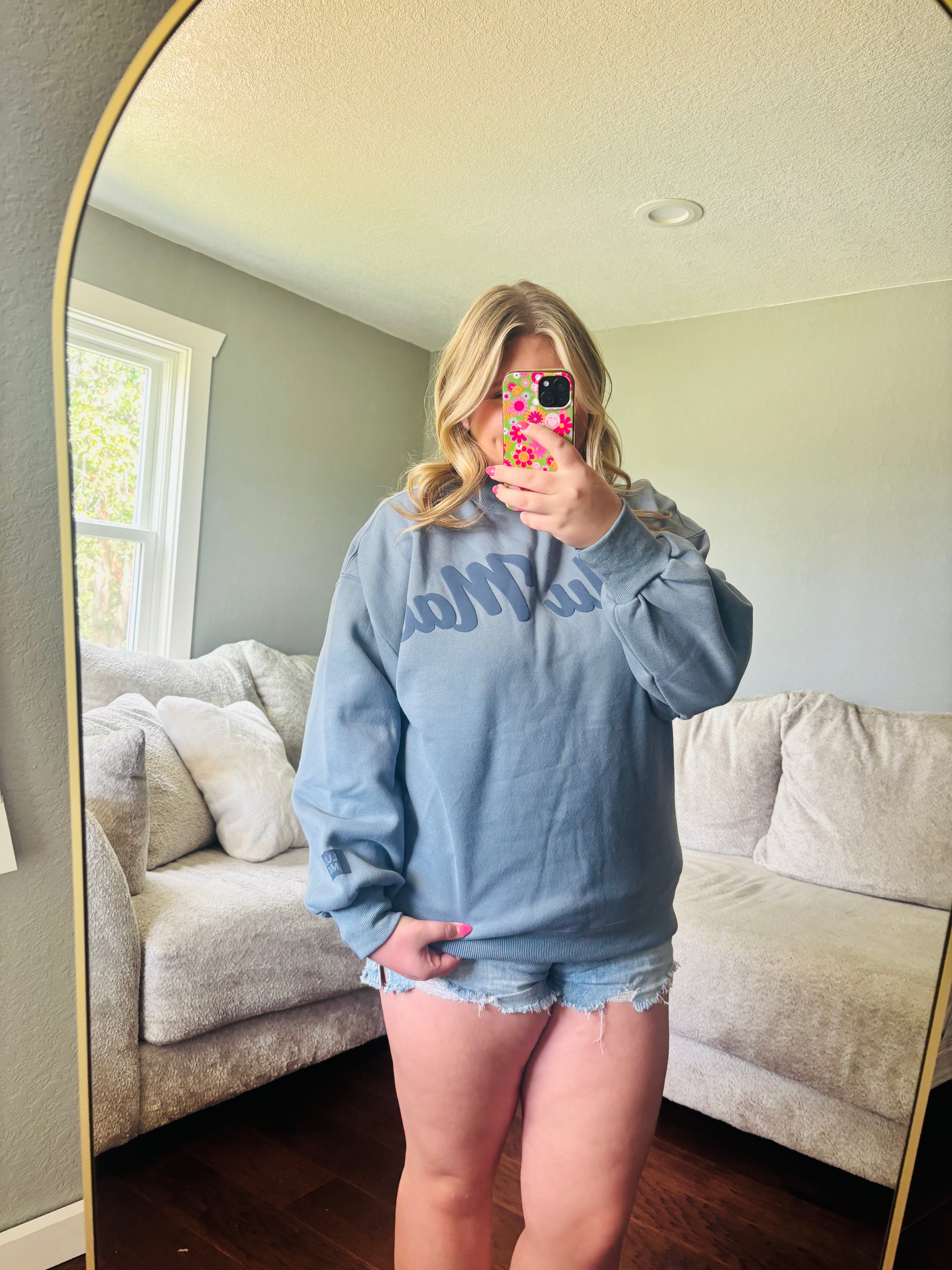A woman takes a mirror selfie in a living room wearing a light blue sweatshirt with a dark blue graphic text across the chest and denim shorts.