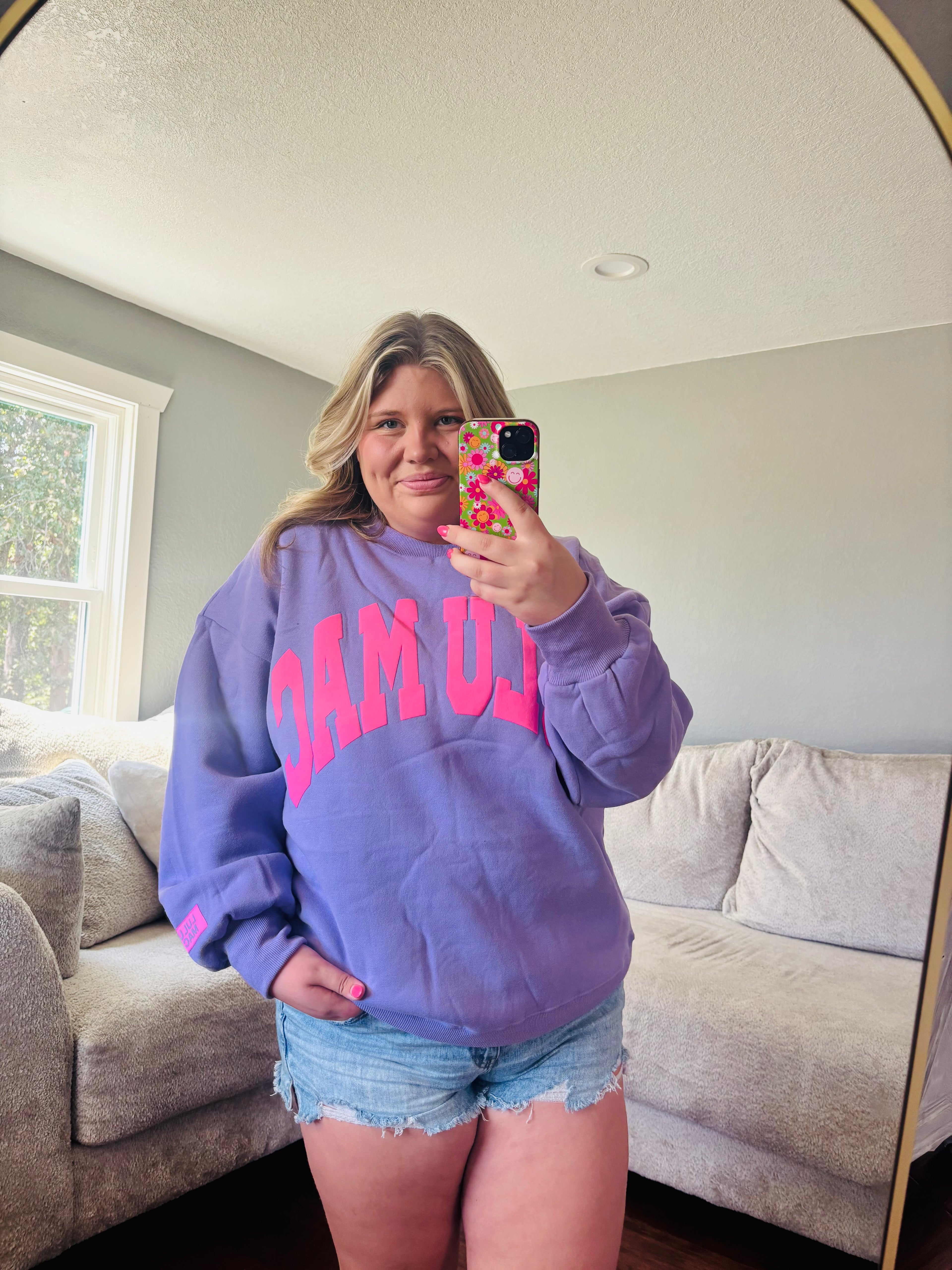 A woman taking a mirror selfie in a living room, wearing a lavender purple sweatshirt with bright pink 'LULU MAC' lettering across the chest and denim shorts.