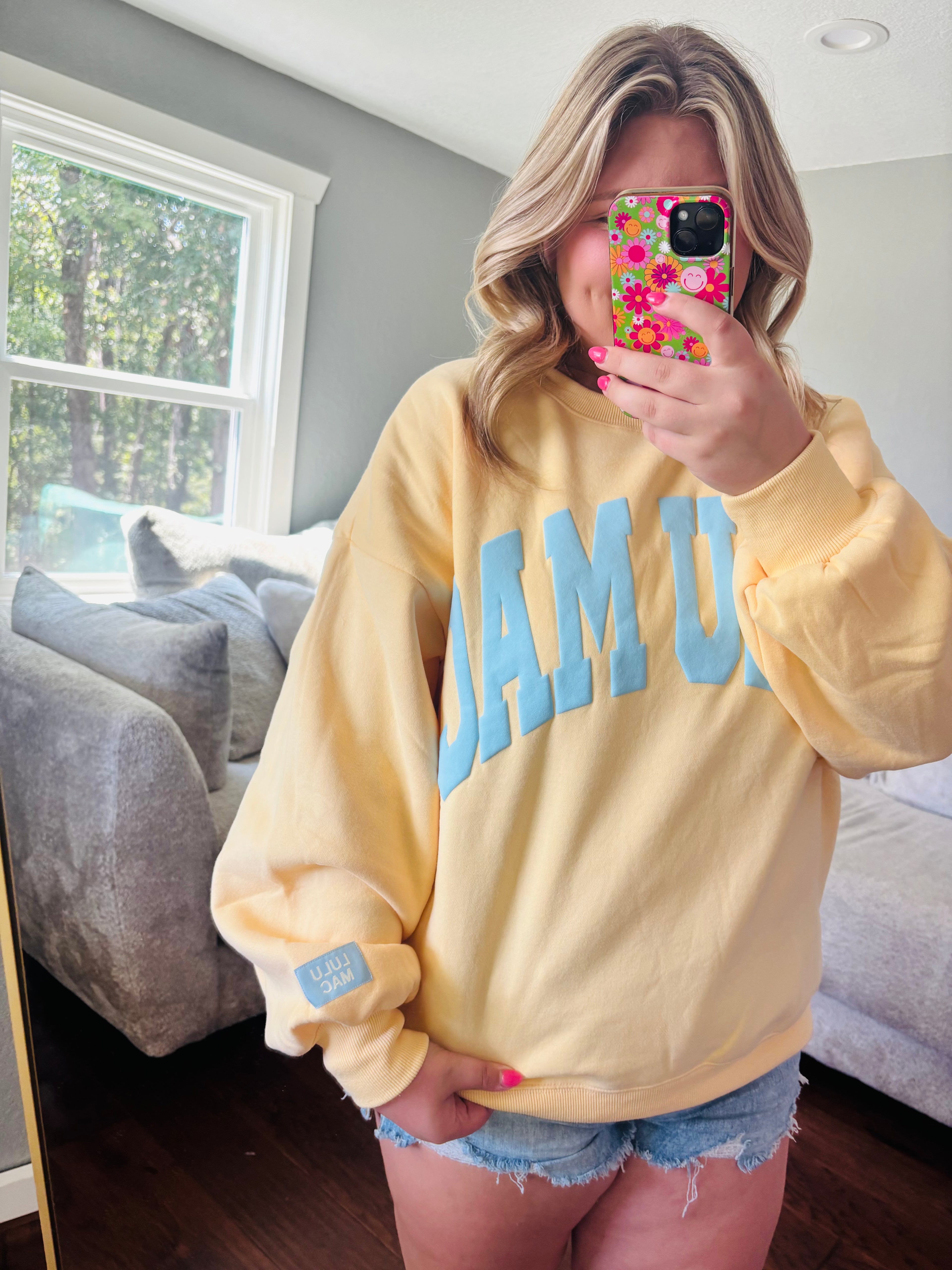 A woman takes a mirror selfie wearing a pale yellow sweatshirt with light blue 'LULU MAC' lettering across the chest, paired with denim shorts in a bedroom setting.