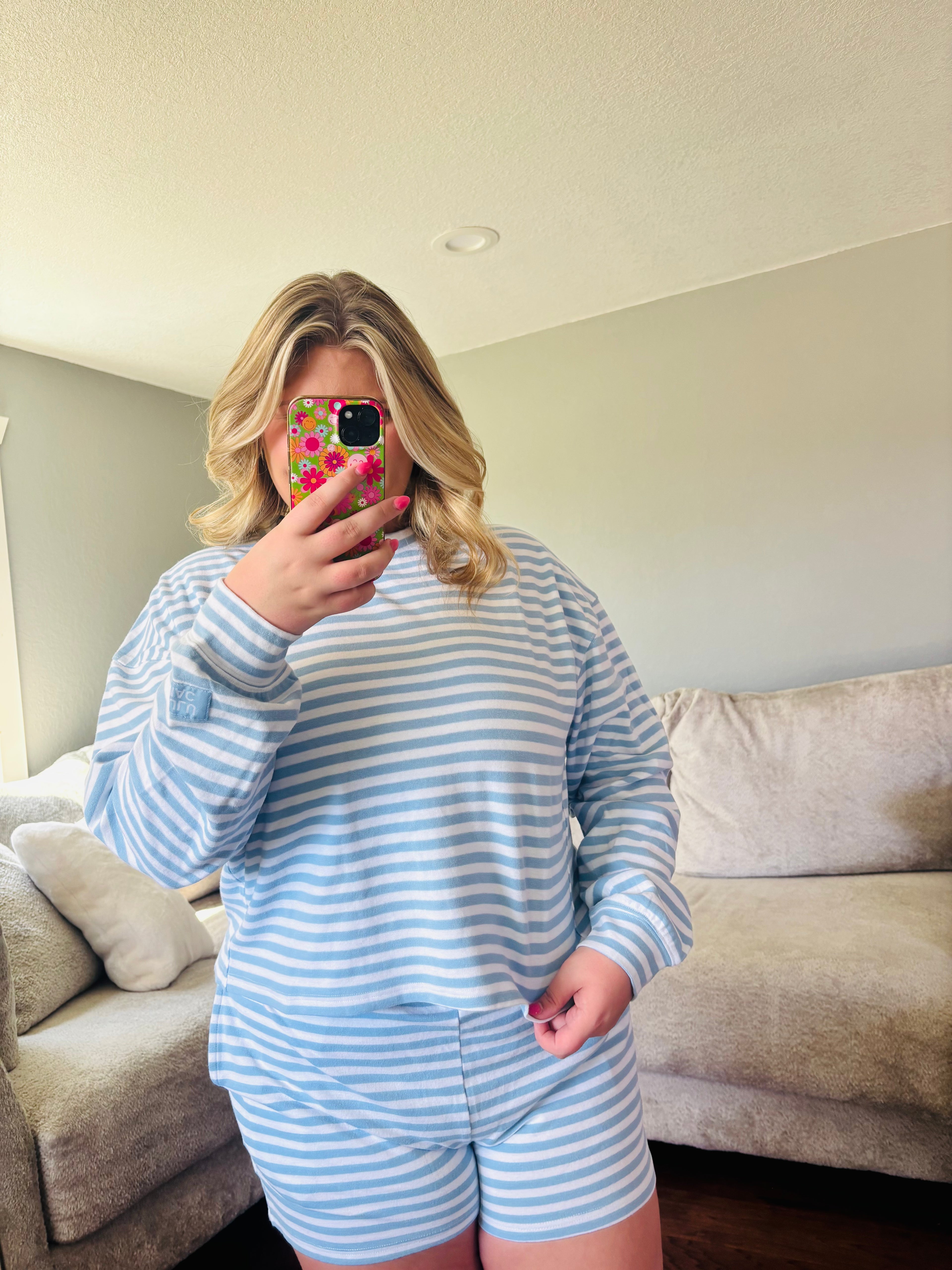 A woman takes a mirror selfie in a living room, wearing a matching light blue and white striped long-sleeve top and shorts set.