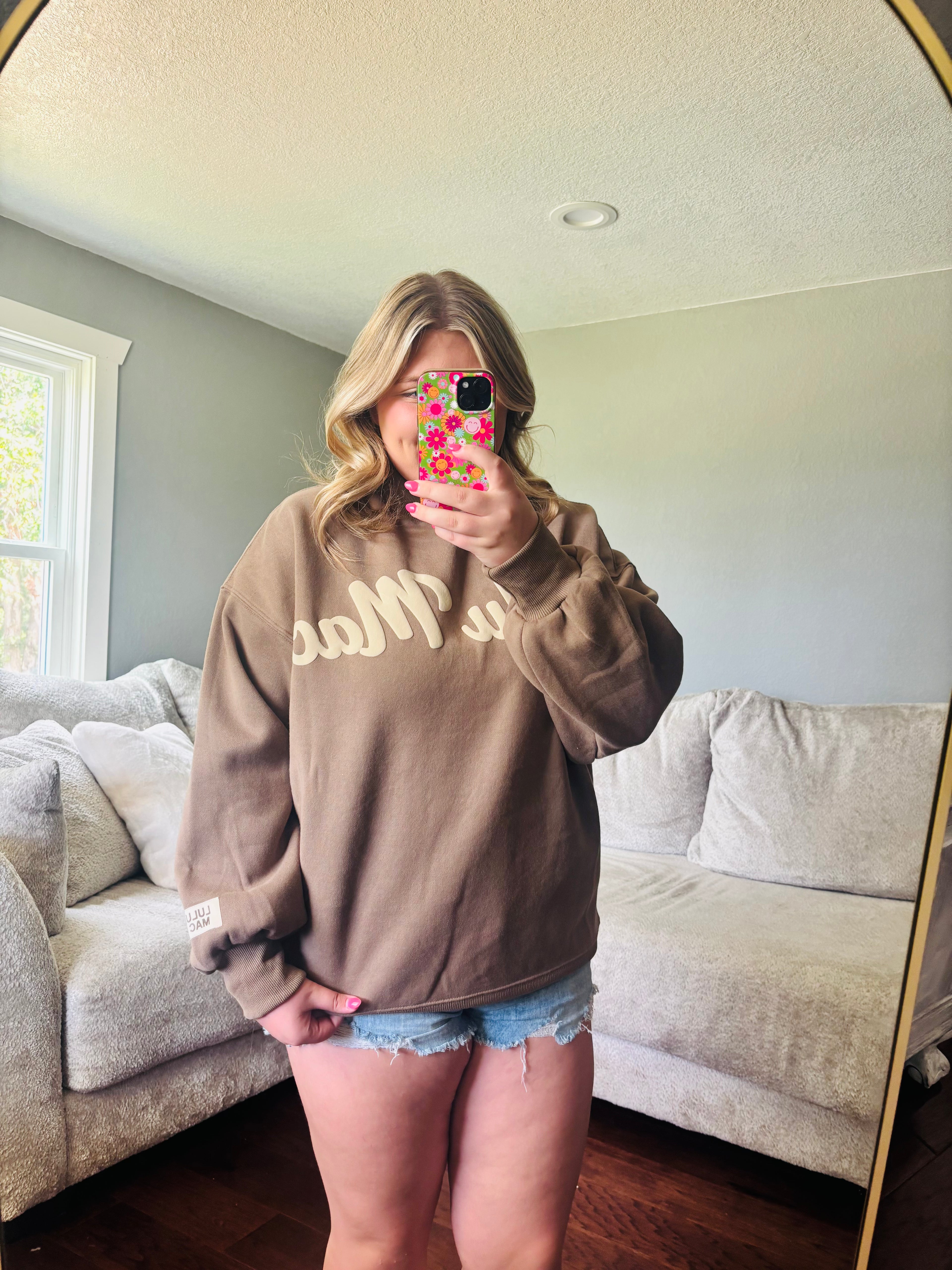 A woman takes a mirror selfie in a living room, wearing an oversized brown sweatshirt with cream lettering that reads 'LULU MAC' and denim shorts.