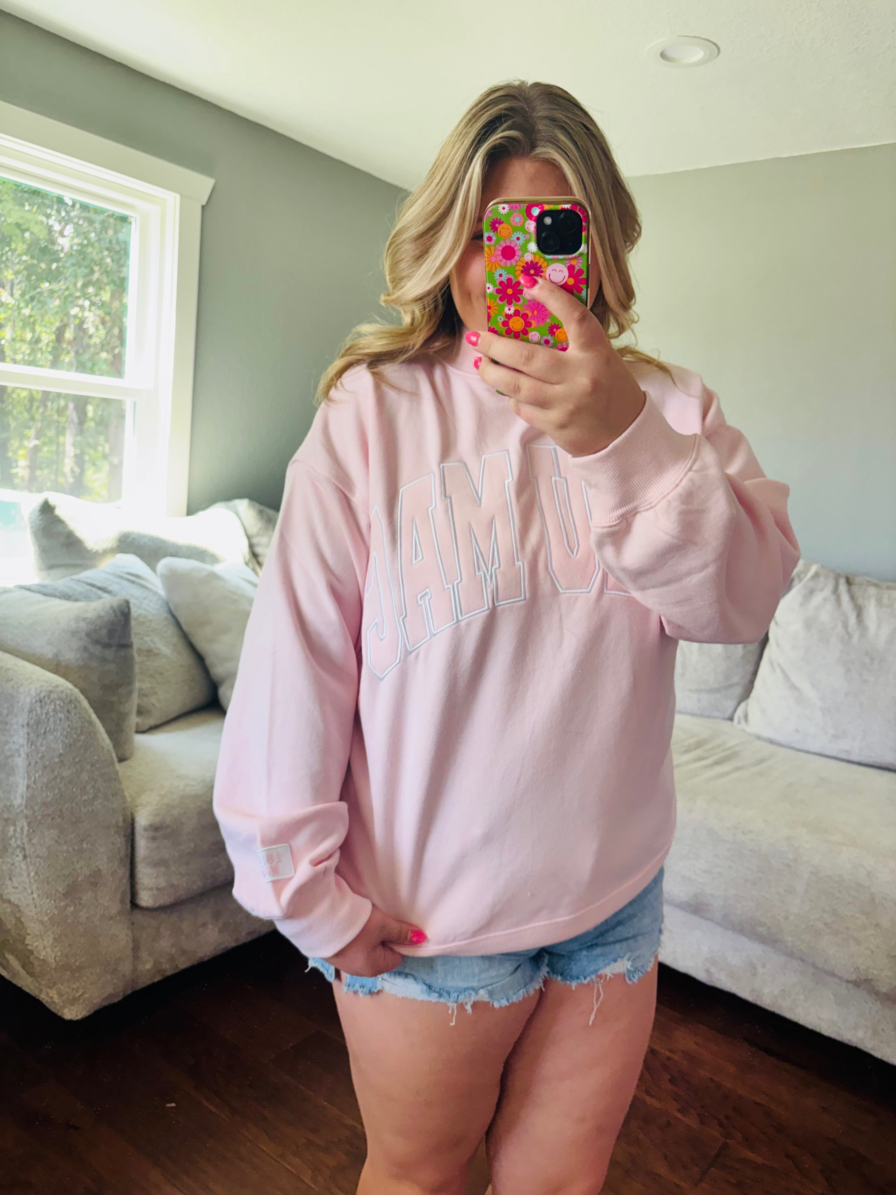 A woman takes a mirror selfie in a living room wearing a light pink crewneck sweatshirt with 'LULU MAC' printed in light blue outline letters across the chest, paired with denim shorts.