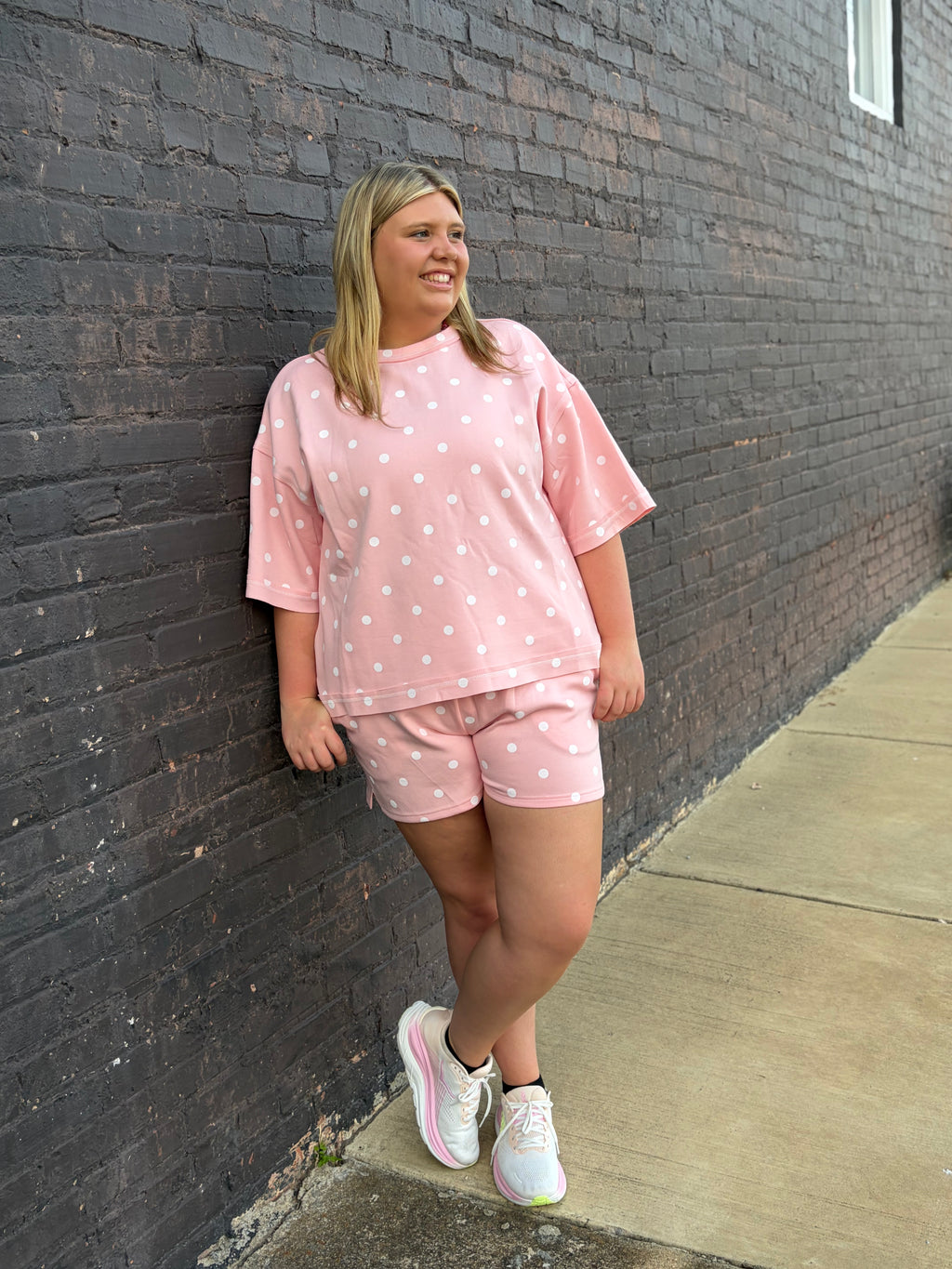 A woman stands against a dark brick wall wearing a light pink short-sleeve t-shirt and matching shorts with white polka dots, paired with white sneakers.