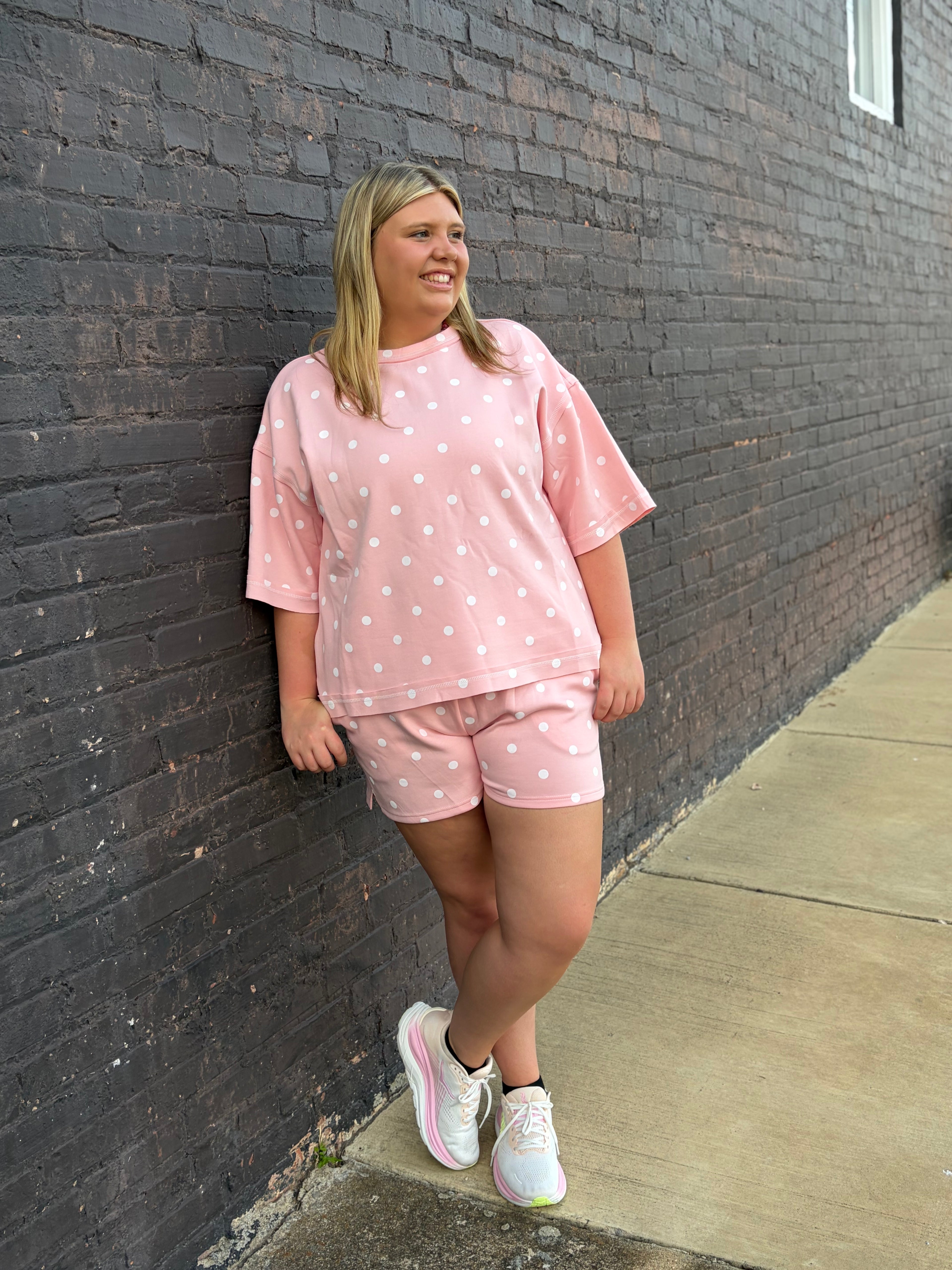 A woman stands against a dark brick wall wearing a light pink short-sleeve t-shirt and matching shorts with white polka dots, paired with white sneakers.