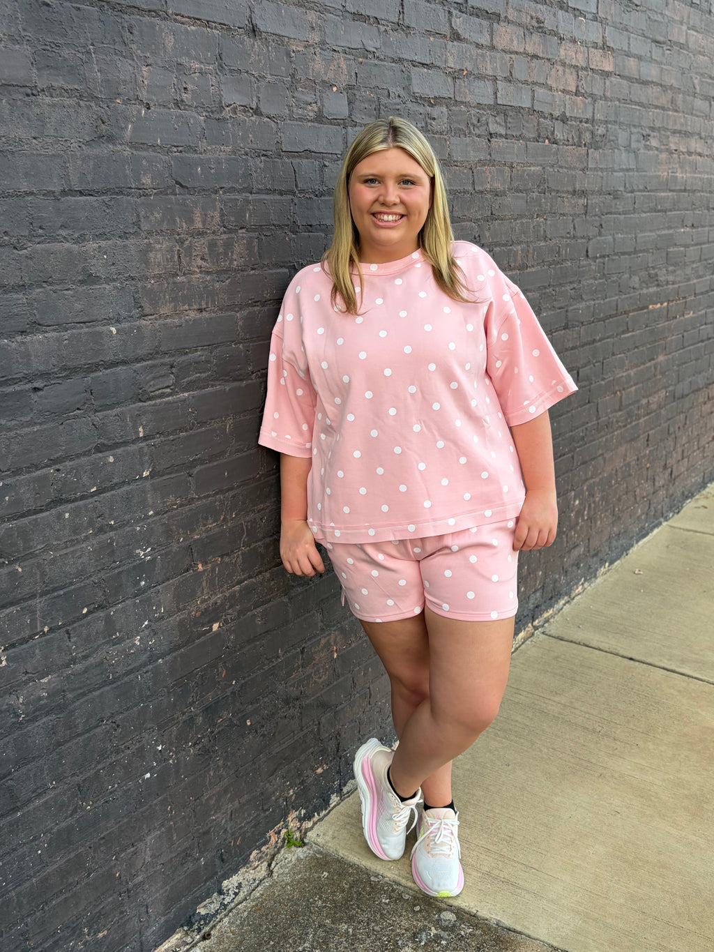 A woman stands against a dark brick wall wearing a light pink short-sleeve t-shirt and matching shorts with white polka dots, paired with white sneakers.