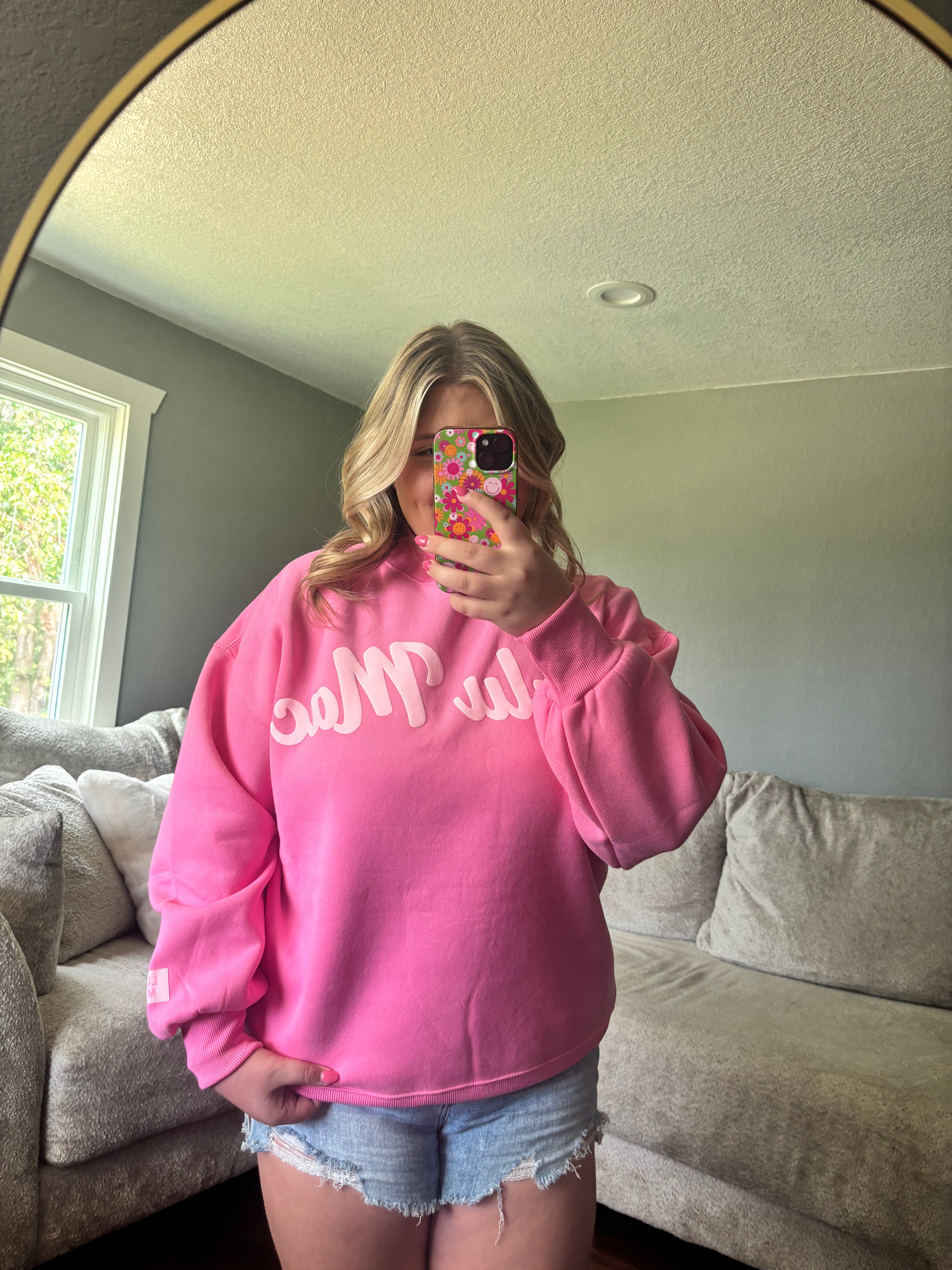 A woman takes a mirror selfie in a living room, wearing a bright pink sweatshirt with white script lettering that reads 'LULU MAC' and light blue denim shorts.