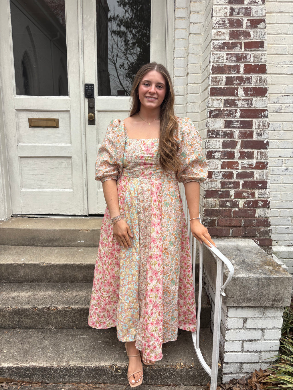 Woman in a floral dress standing on a set of stairs with a white door and brick wall in the background.