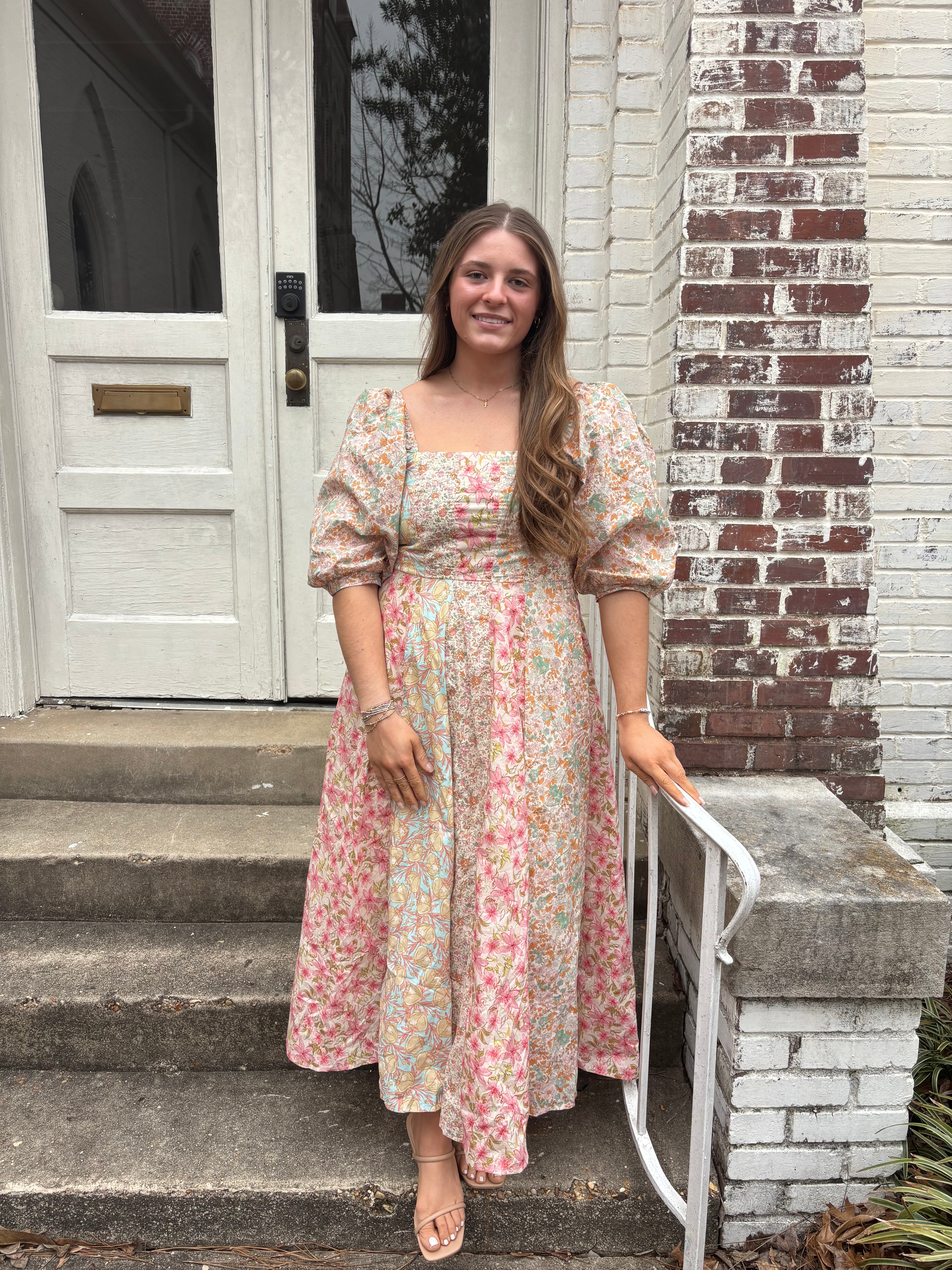 Woman in a floral dress standing on a set of stairs with a white door and brick wall in the background.