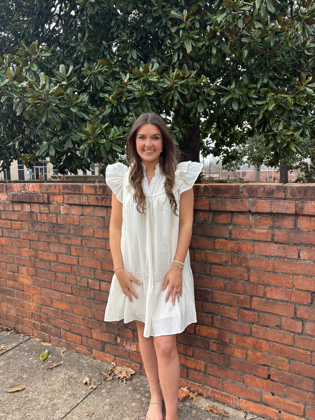 Woman in a white dress standing against a brick wall with trees in the background