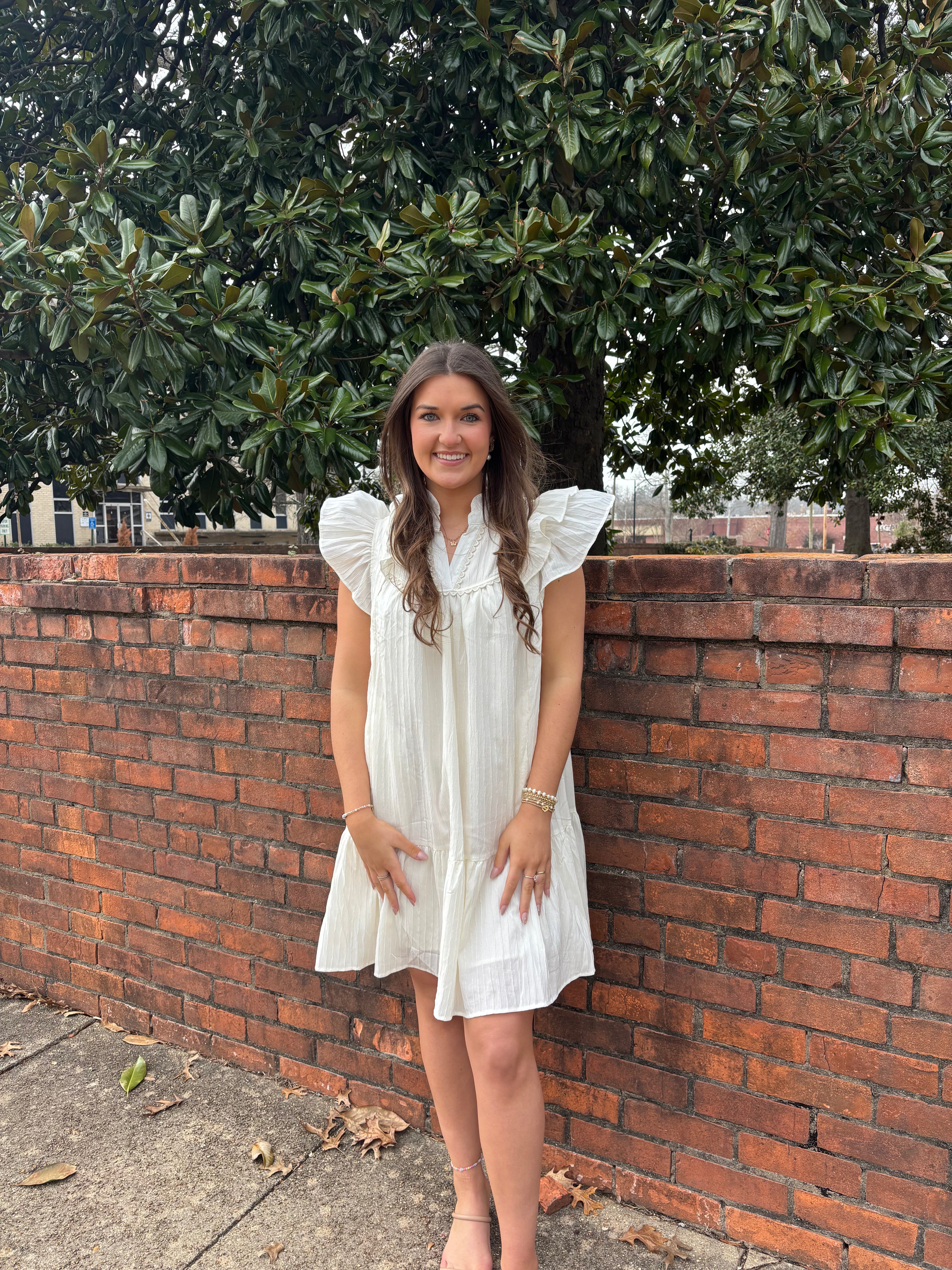 Woman in a white dress standing against a brick wall with trees in the background