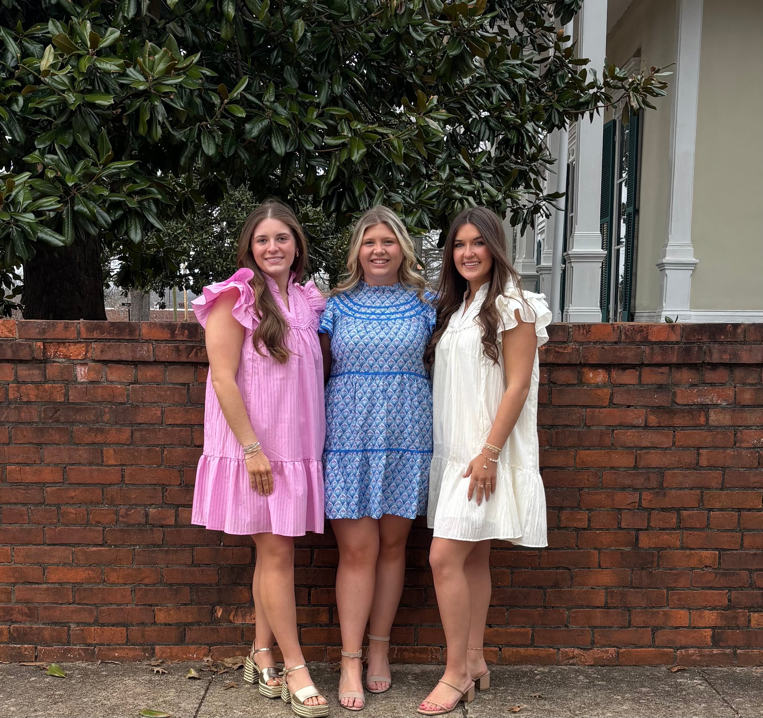 Three women standing together in front of a brick wall with a tree in the background