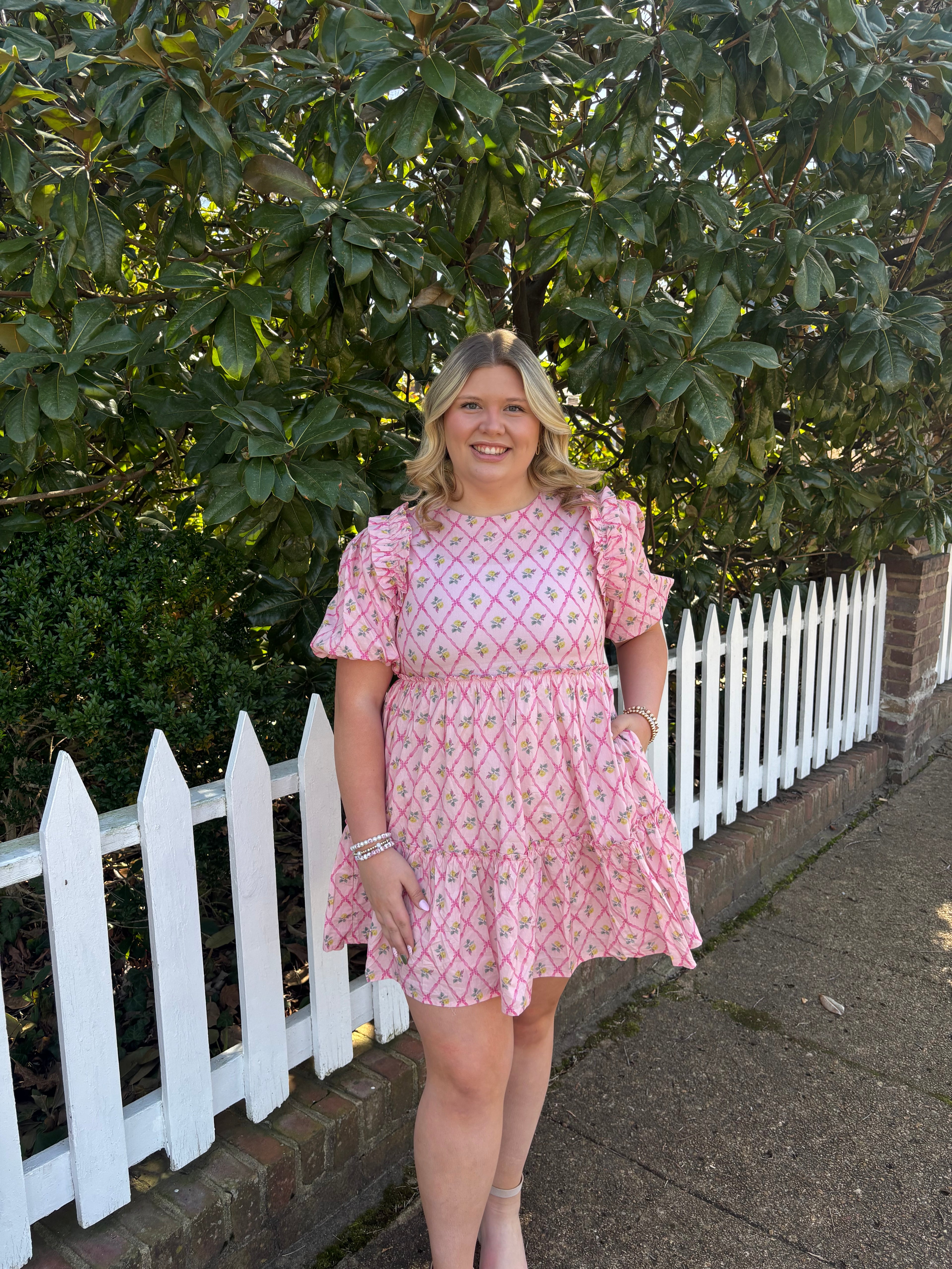 Woman in a pink dress standing next to a white picket fence with greenery and pavement in the background