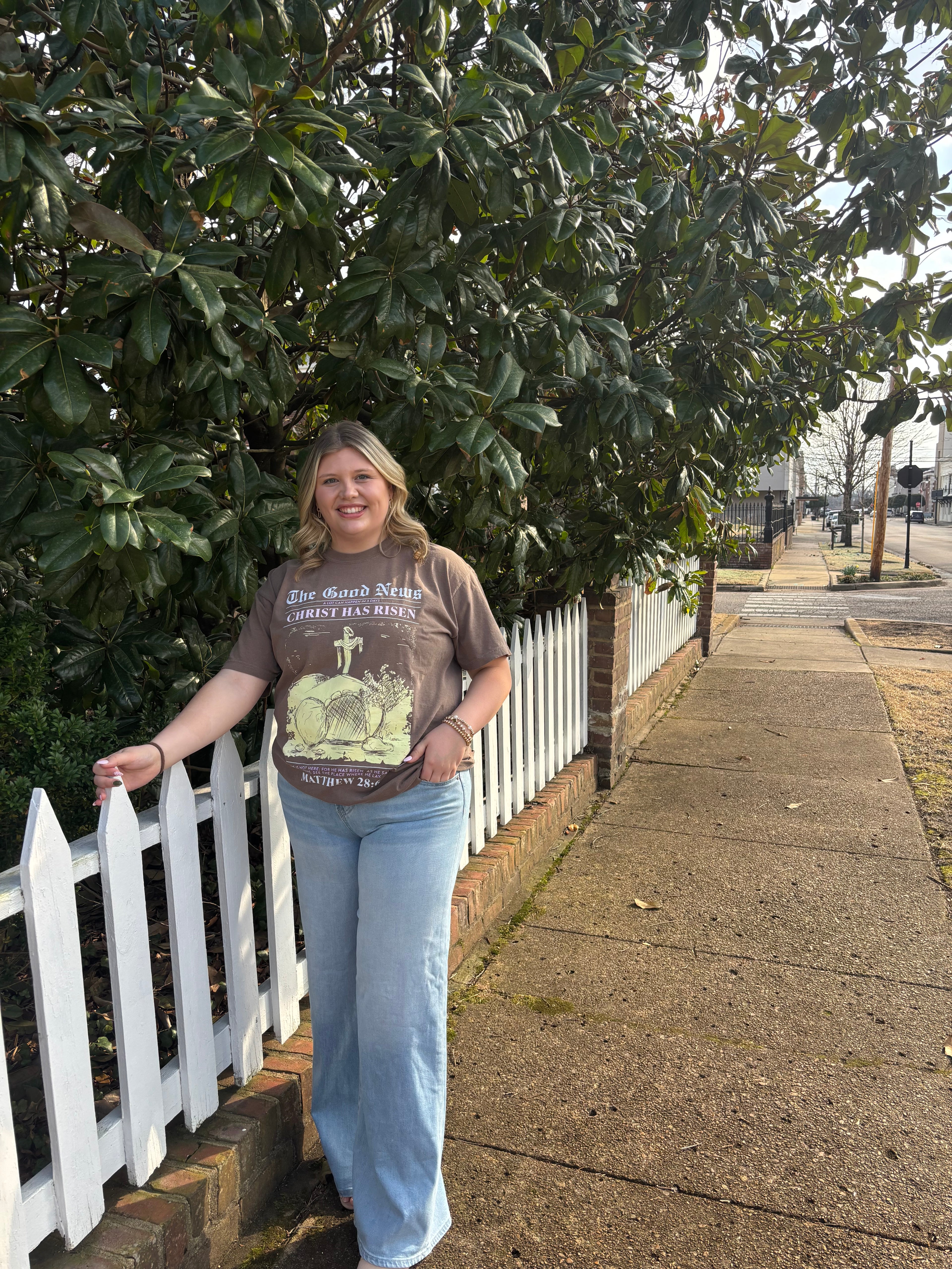 A woman with light washed jeans and a brown t-shirt standing outside next to a white picket fence 